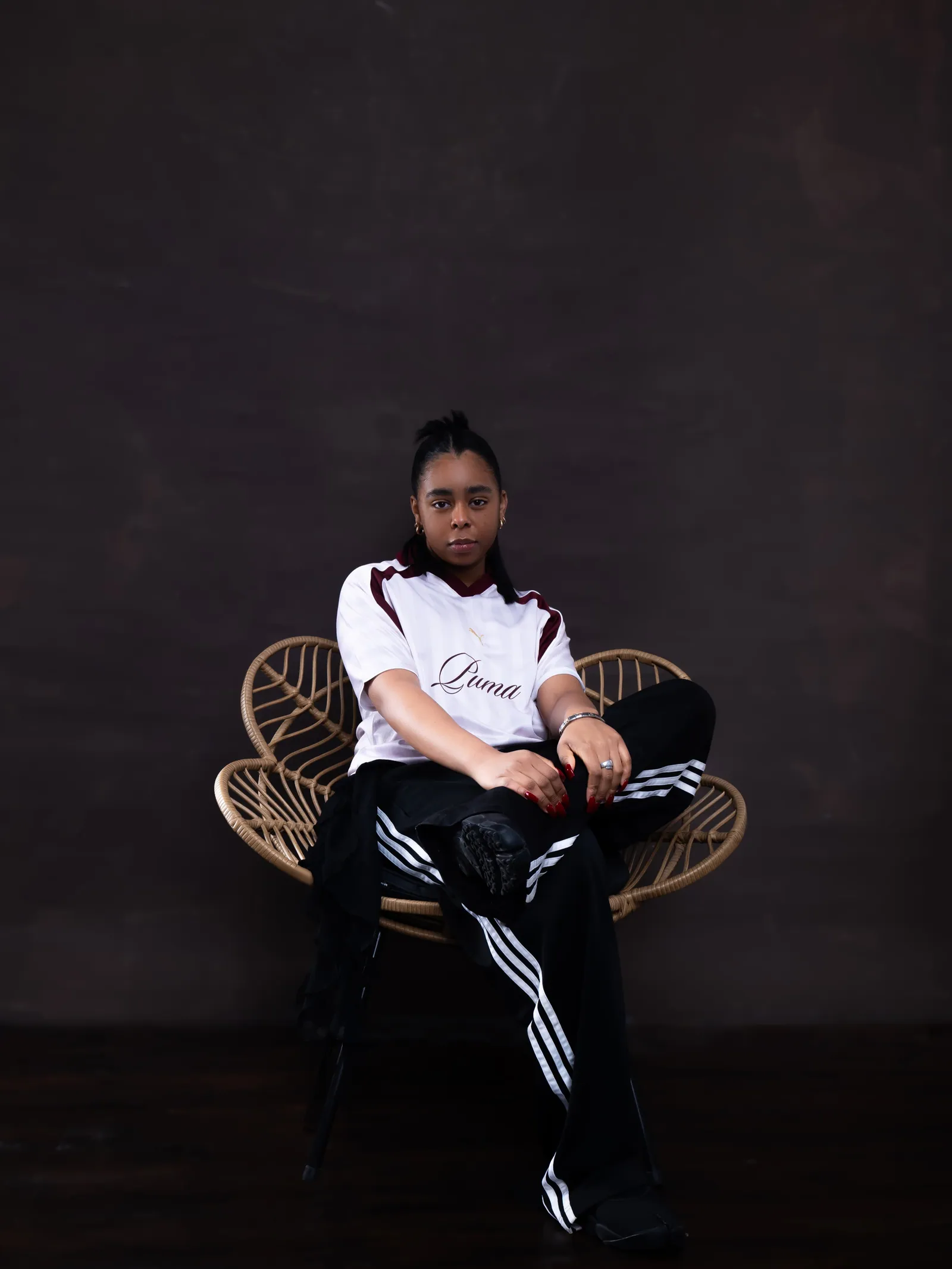 A woman in a white Puma football shirt and black striped track trousers seated in a rattan chair against a dark backdrop