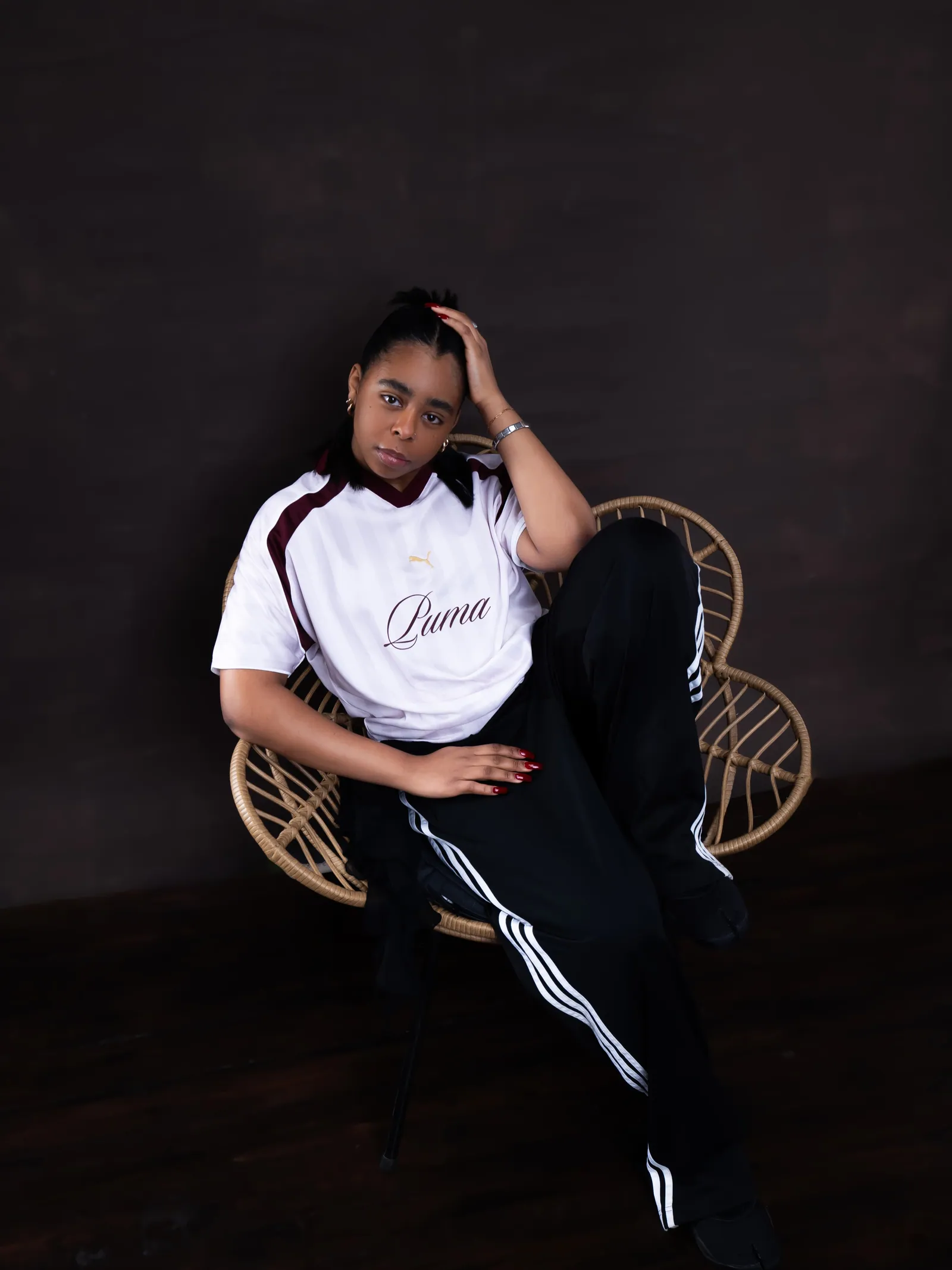A woman in a white Puma shirt and black striped trousers lounging in a rattan chair, hand on her head, dark backdrop
