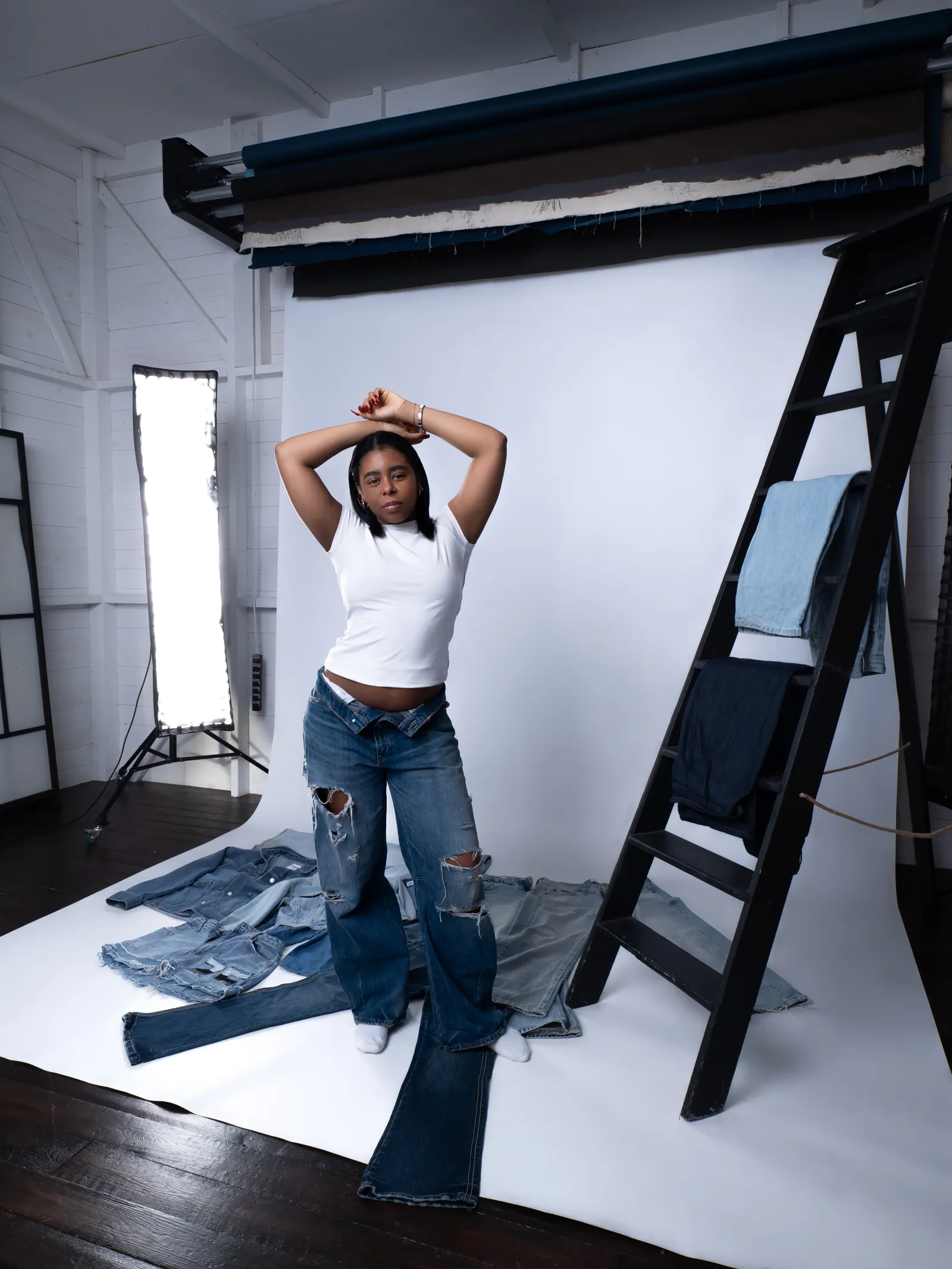 A woman in a white tee and ripped jeans posing with arms overhead on a white cyclorama, denim laid out, light stand beside