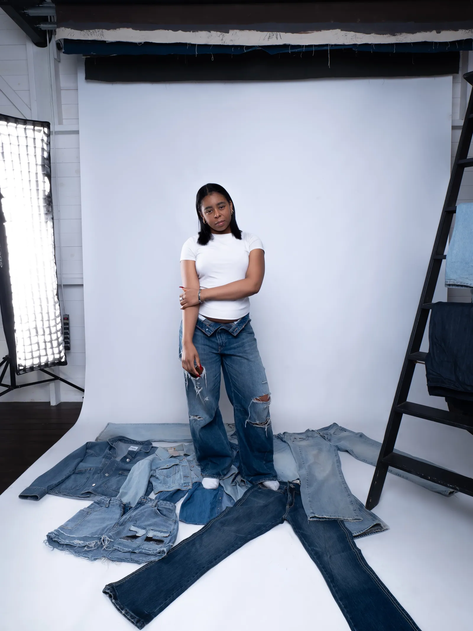 A woman in a white tee and ripped jeans standing on a white backdrop among laid-out denim jackets and jeans, softbox left