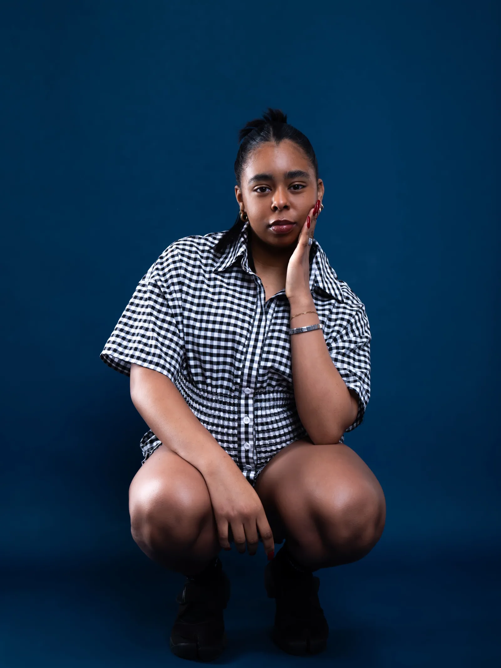 A woman in a black and white gingham shirt crouching with her hand on her cheek against a deep blue backdrop