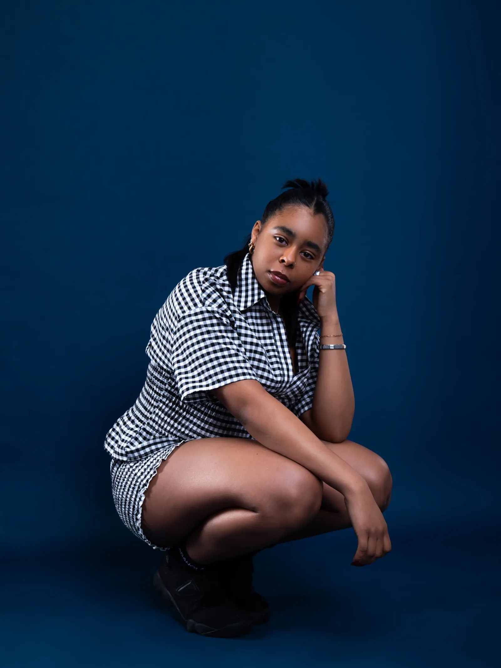 A woman in a gingham shirt and shorts crouching with her head on her hand against a deep blue studio backdrop