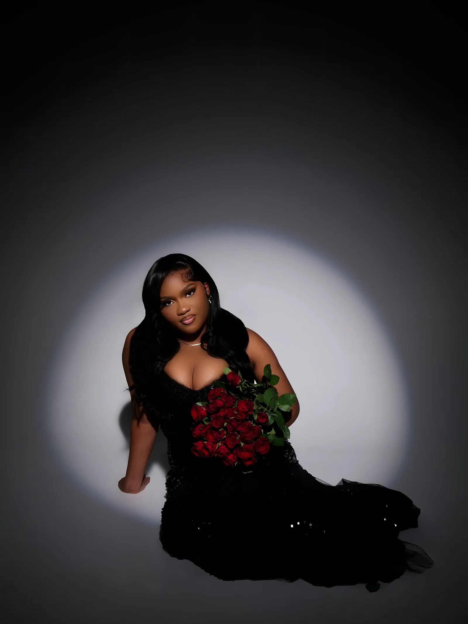 Woman in a black sequinned gown seated in a spotlight on a grey floor, holding a bouquet of red roses