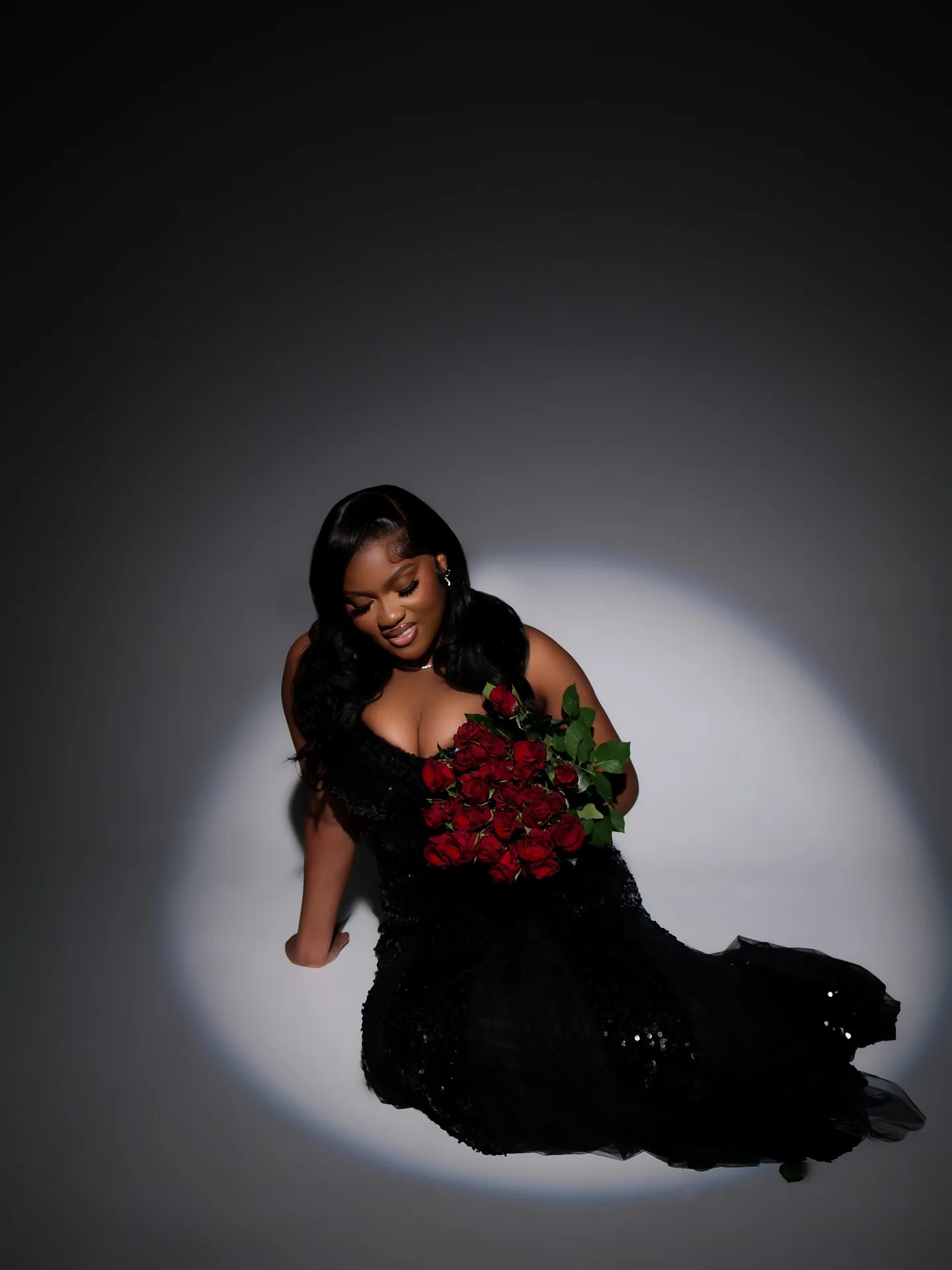 Woman in a black gown looking down and smiling, holding red roses, seated in a pool of white light