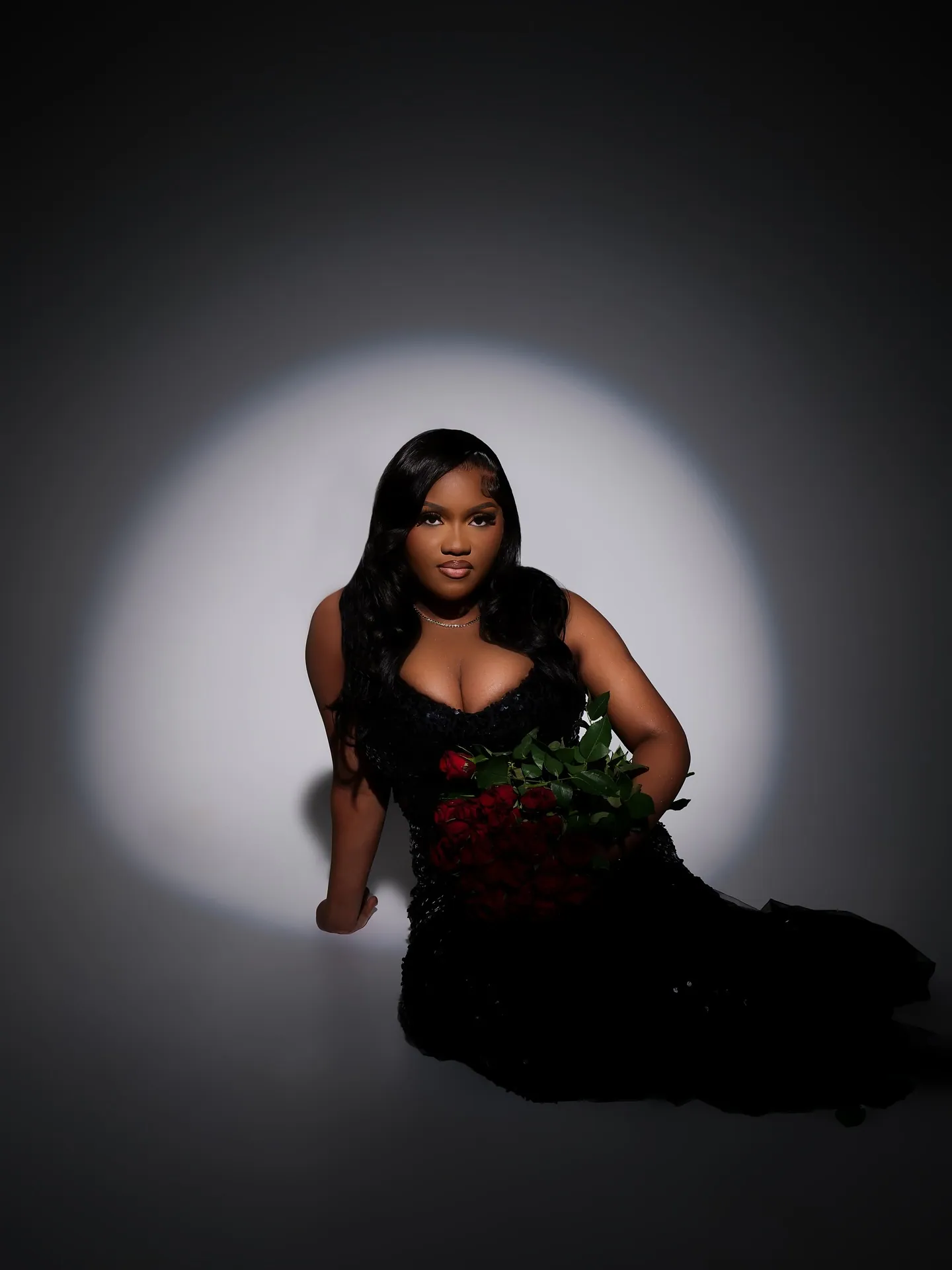 Woman in a black gown looking at the camera, red roses in her arm, seated in a spotlight on a dark floor