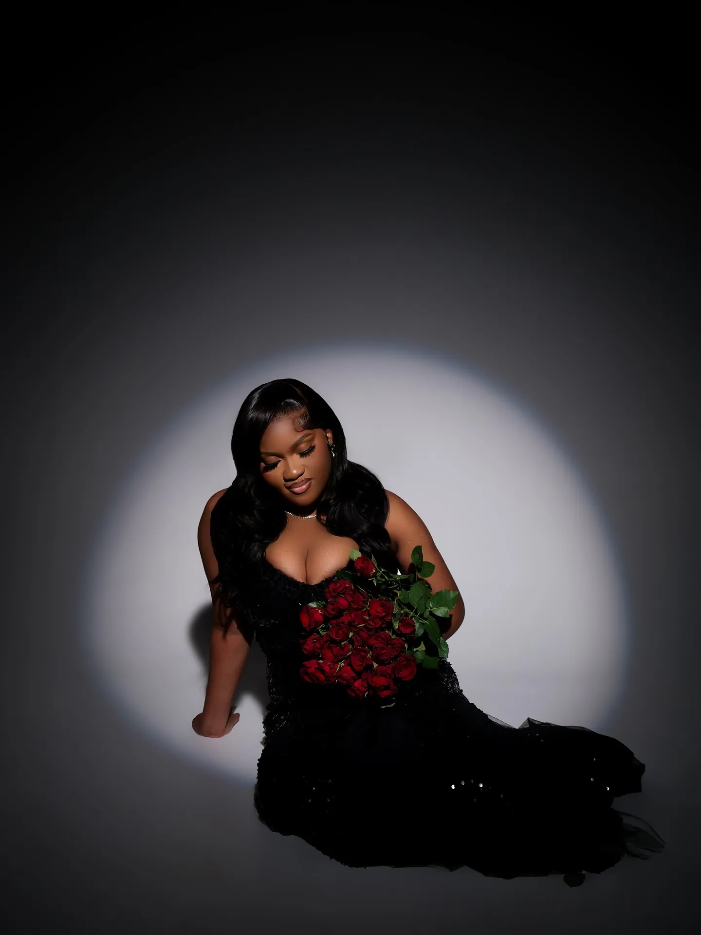 Woman in a black sequinned gown with long hair looking down at a red rose bouquet, white spotlight circle