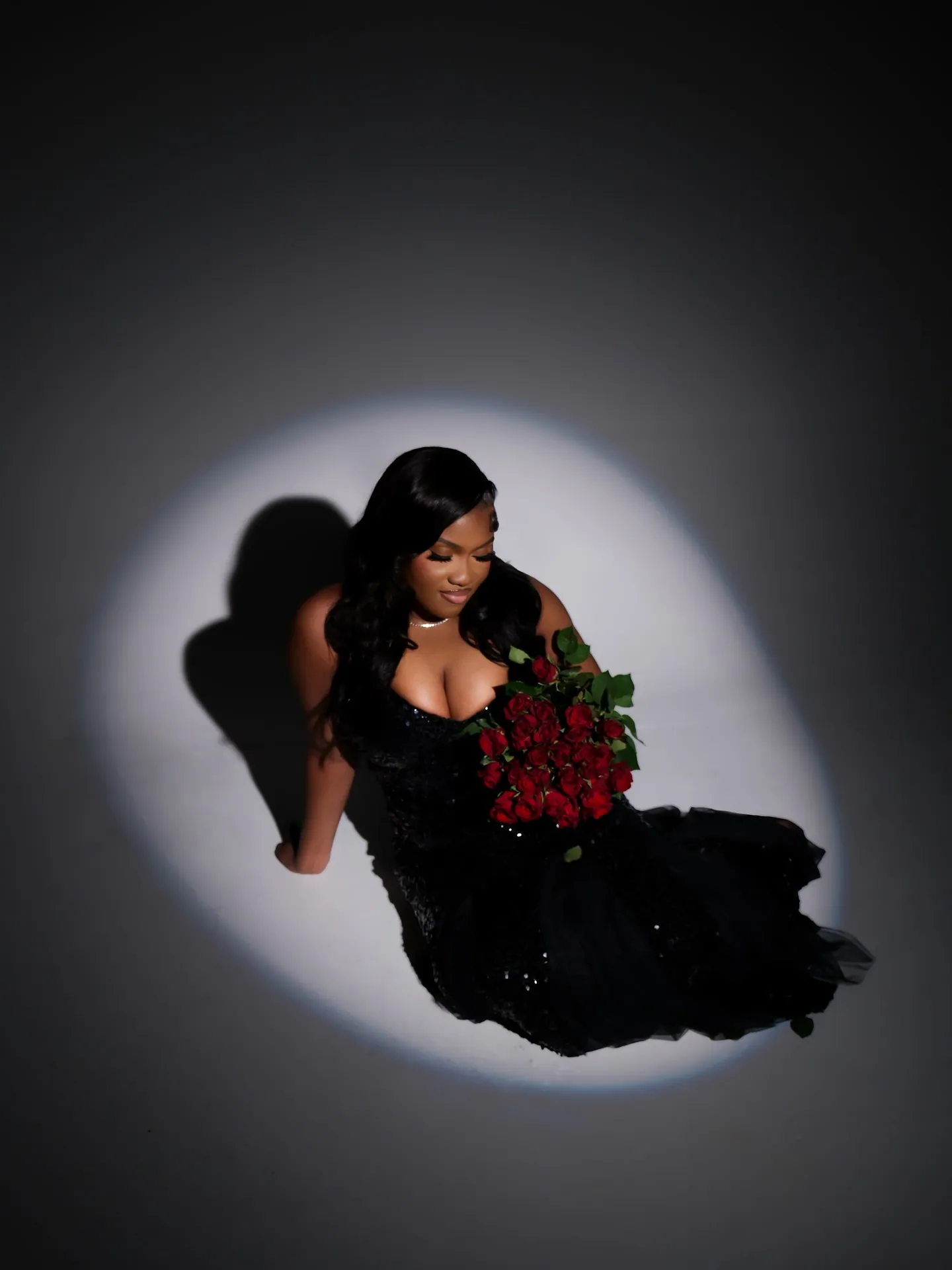 Overhead view of a woman in a black gown holding red roses, her shadow cast in a white spotlight