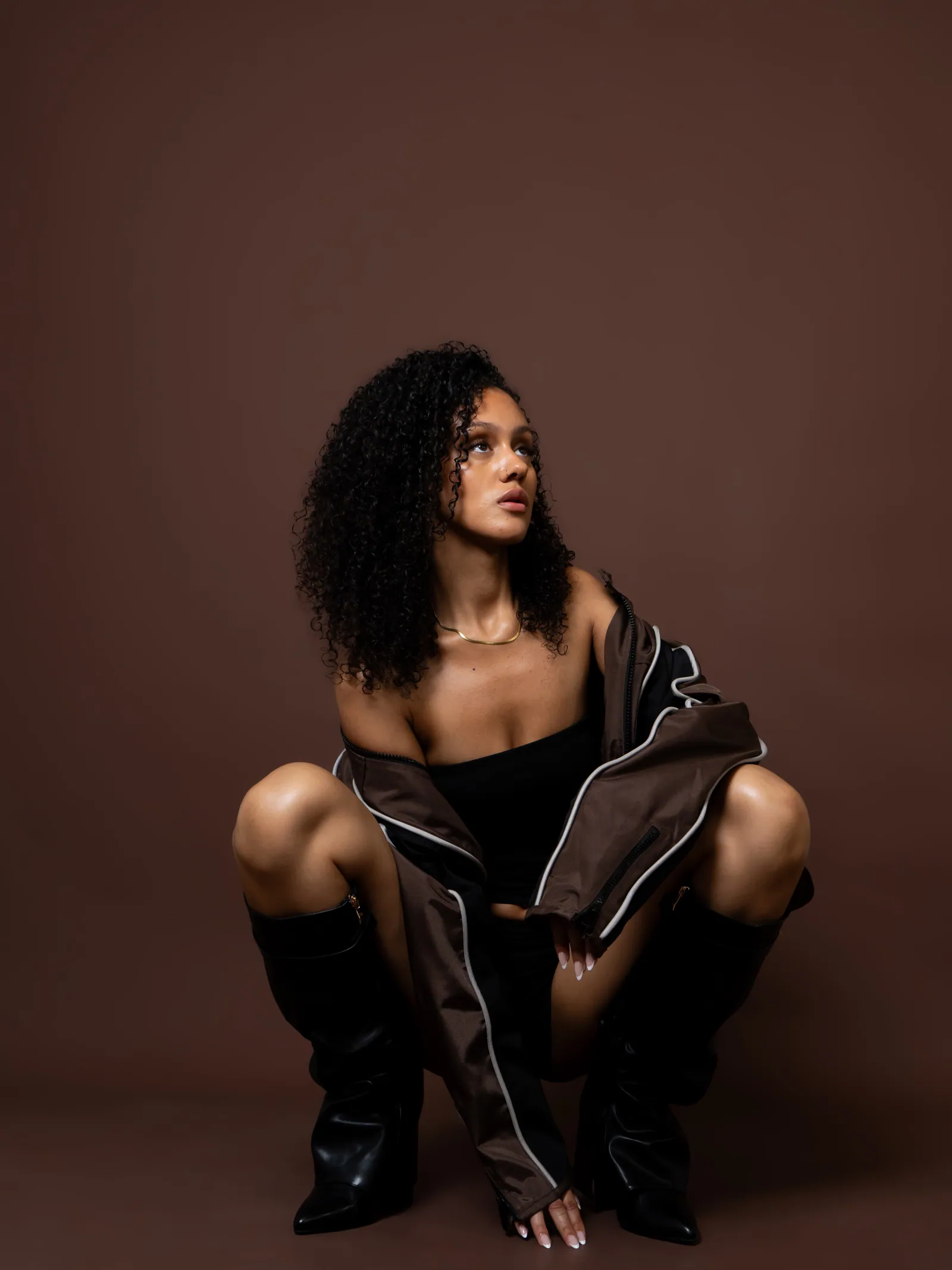Woman with curly hair crouching in black boots and a brown jacket slipping off her shoulders, brown backdrop