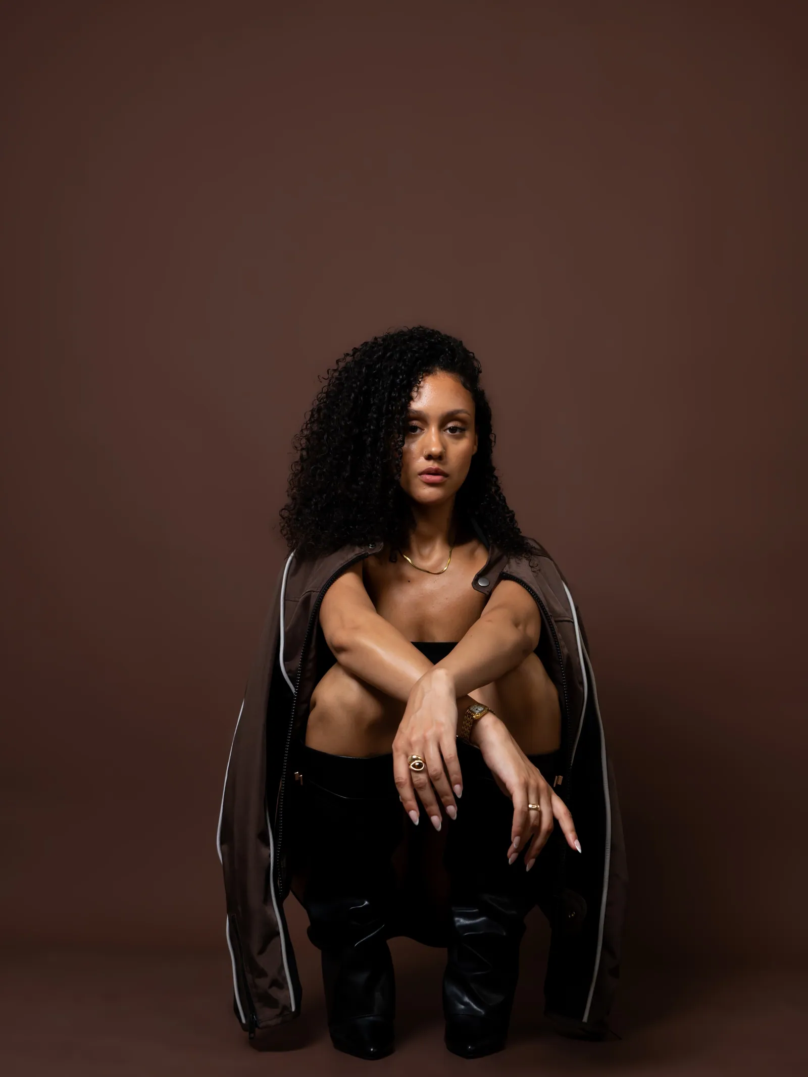 Woman with curly hair sitting crouched, arms crossed over her knees, brown jacket draped on her shoulders