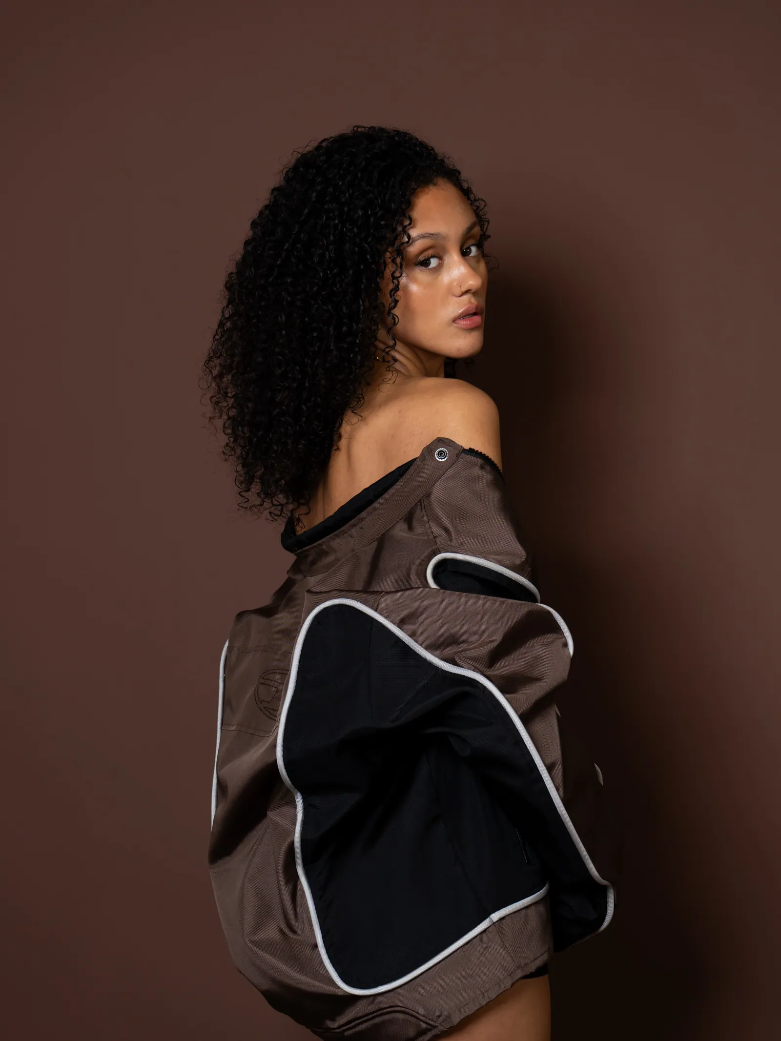 Woman with curly hair looking back over her bare shoulder in an oversized brown jacket with white piping