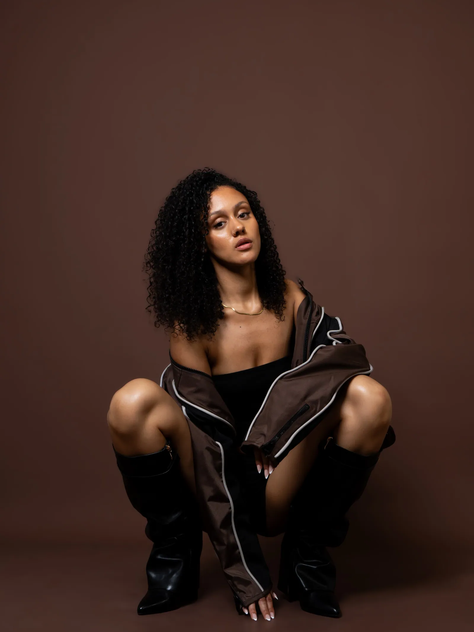 Woman with curly hair crouching and looking at the camera in a brown jacket and black boots, brown backdrop