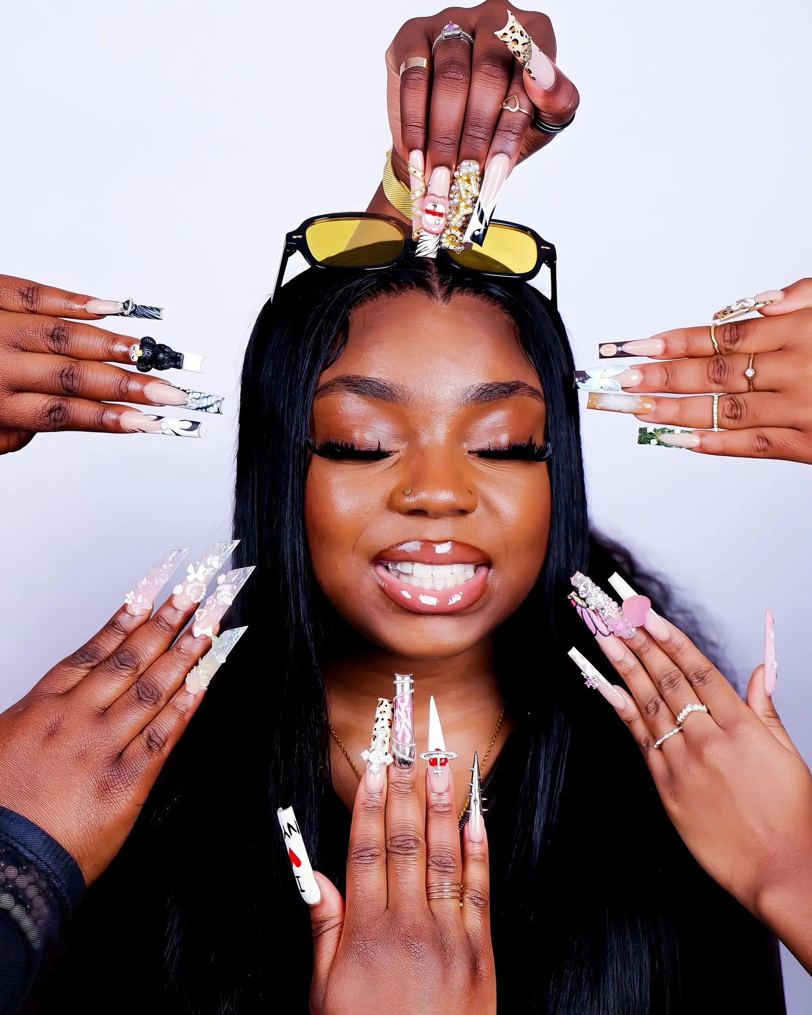 A smiling woman with closed eyes and yellow sunglasses on her head, ringed by hands with long decorated nails, white backdrop
