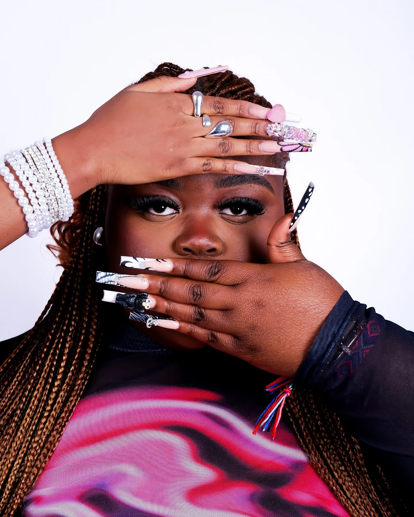 A woman with long braids covering her mouth with both hands, long patterned nails and pearl bracelets, pink swirl top