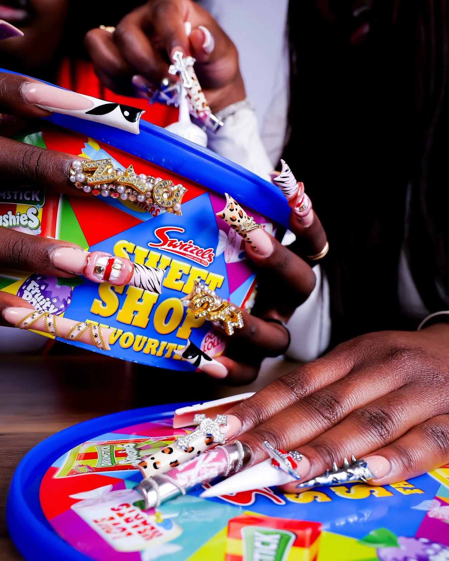 Hands with long jewelled and animal-print nails around a blue Swizzels Sweet Shop tub, bright sweet packaging