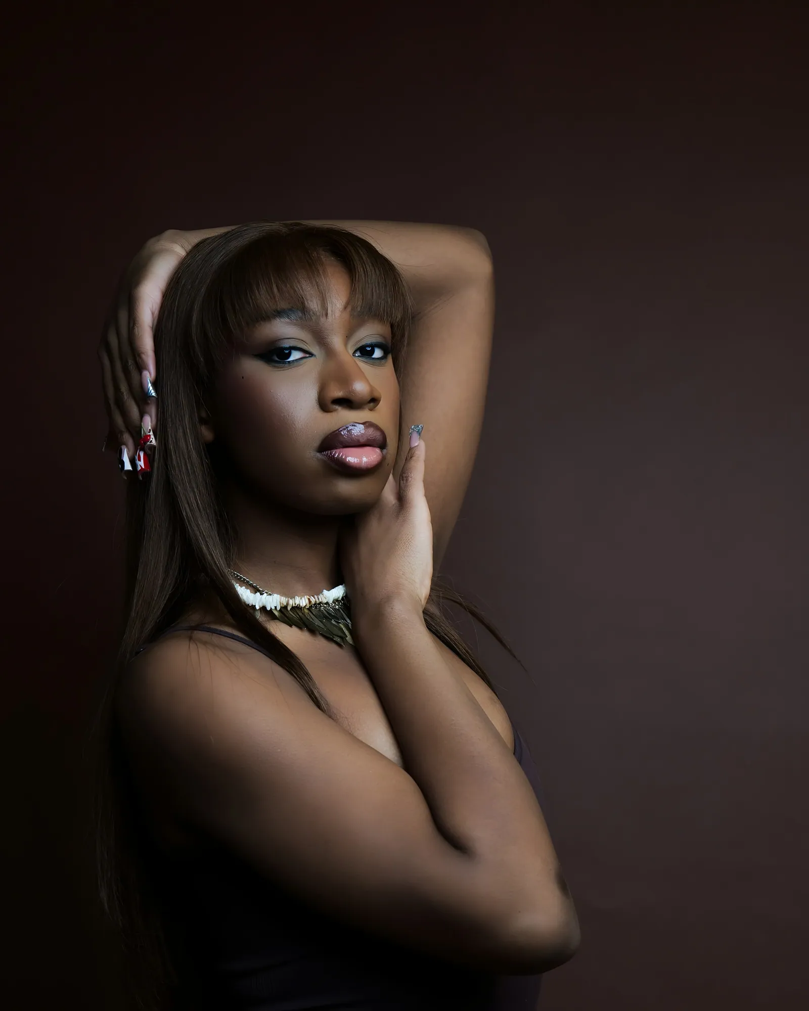 A woman with long hair and a fringe, arm raised over her head, wearing a white bead necklace against a dark brown backdrop