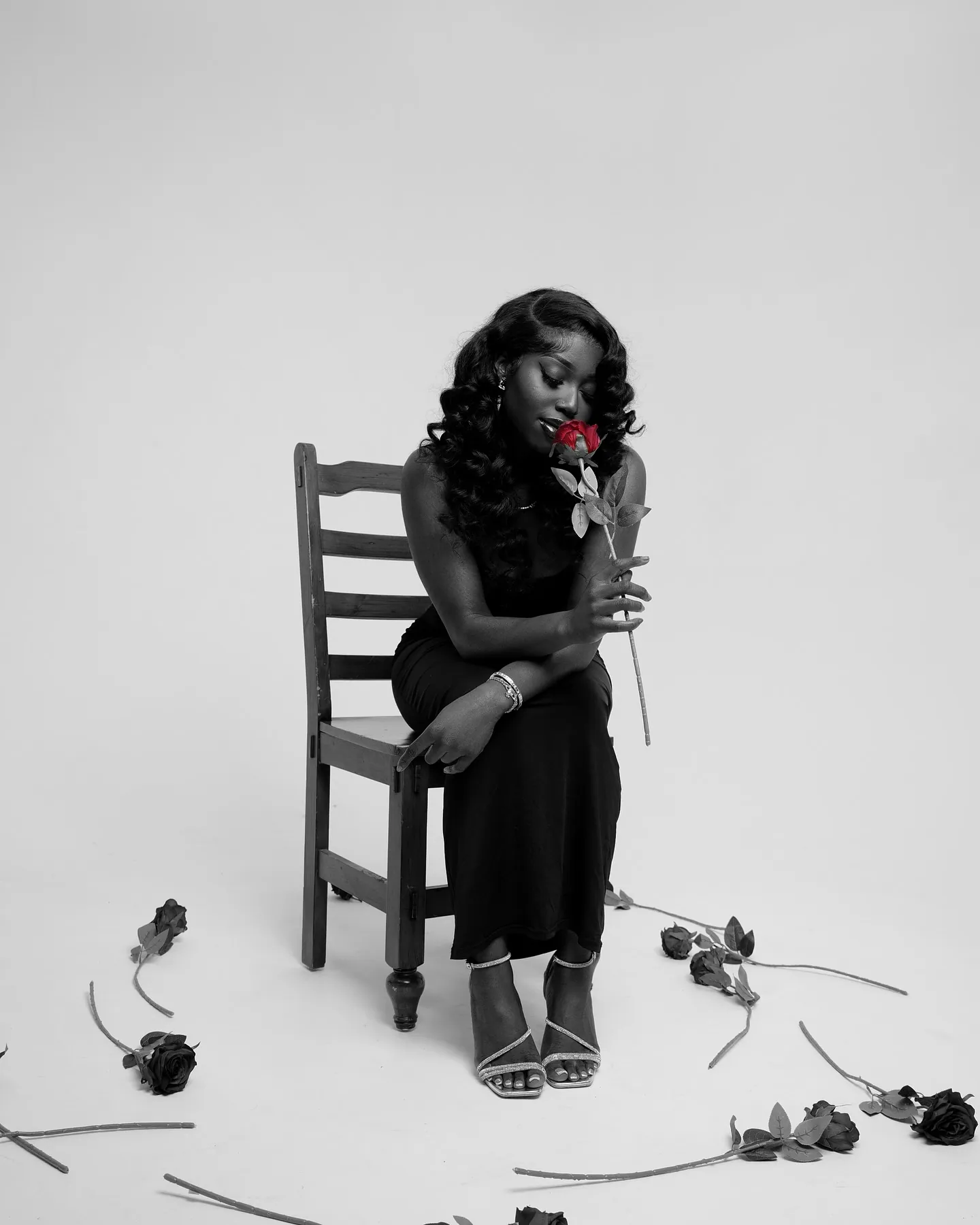 Black and white shot of a woman in a black dress seated on a chair holding a single red rose, roses scattered on the floor