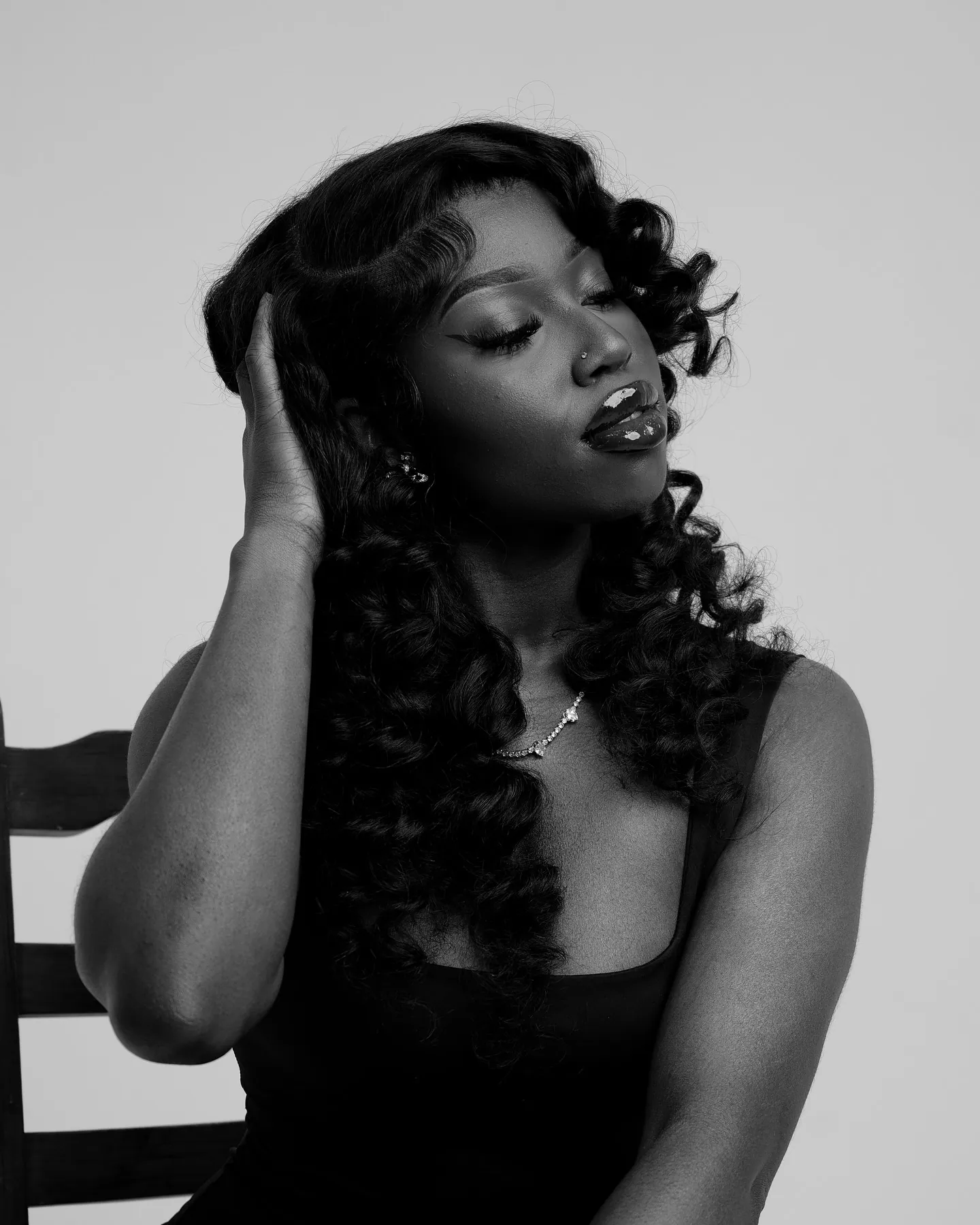 Black and white portrait of a woman with curled hair, eyes closed, one hand in her hair, wearing a black top