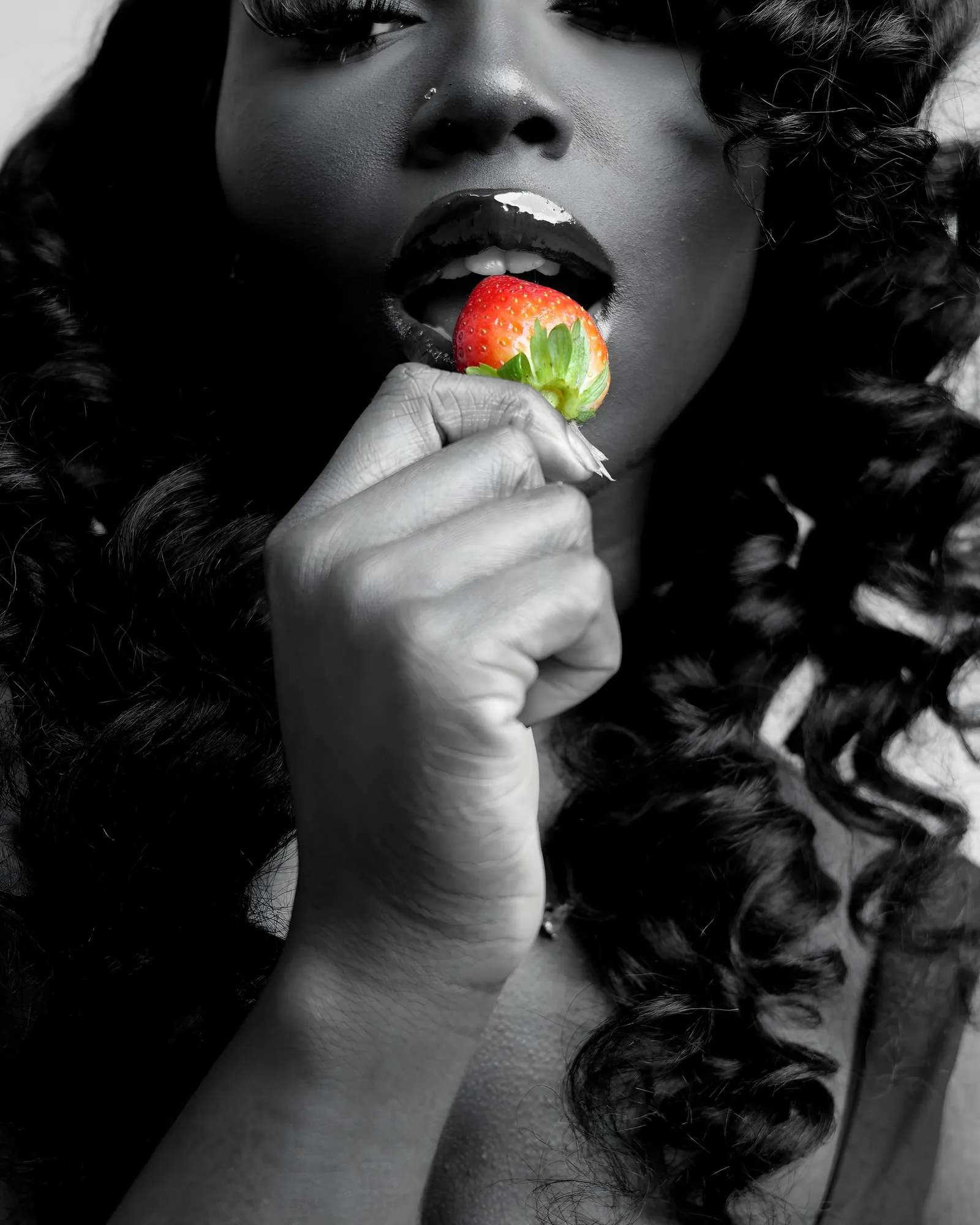 Black and white close-up of a woman with curled hair biting a strawberry, which is the only colour in the frame