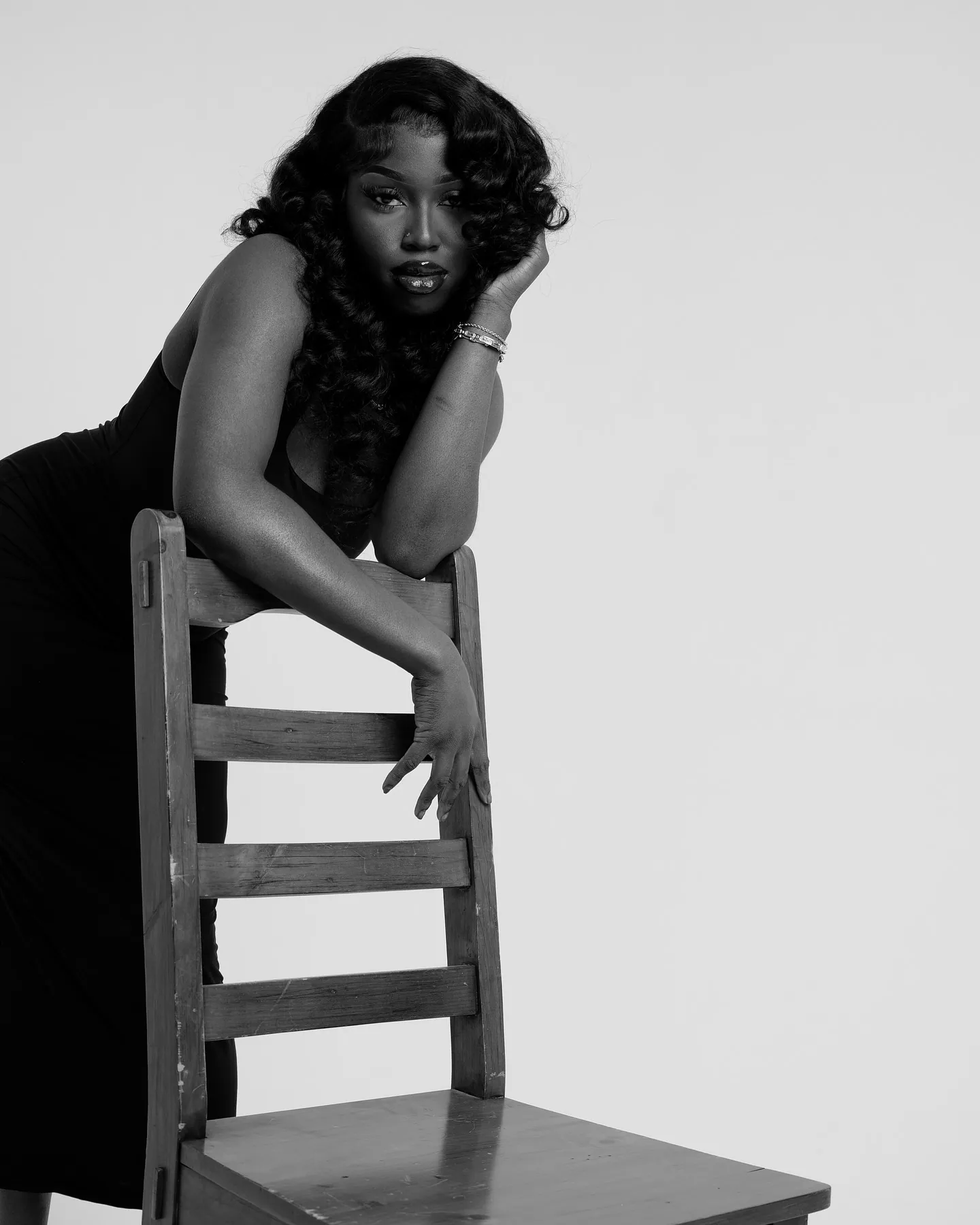 Black and white shot of a woman in a black dress leaning over the back of a wooden chair, head on her hand