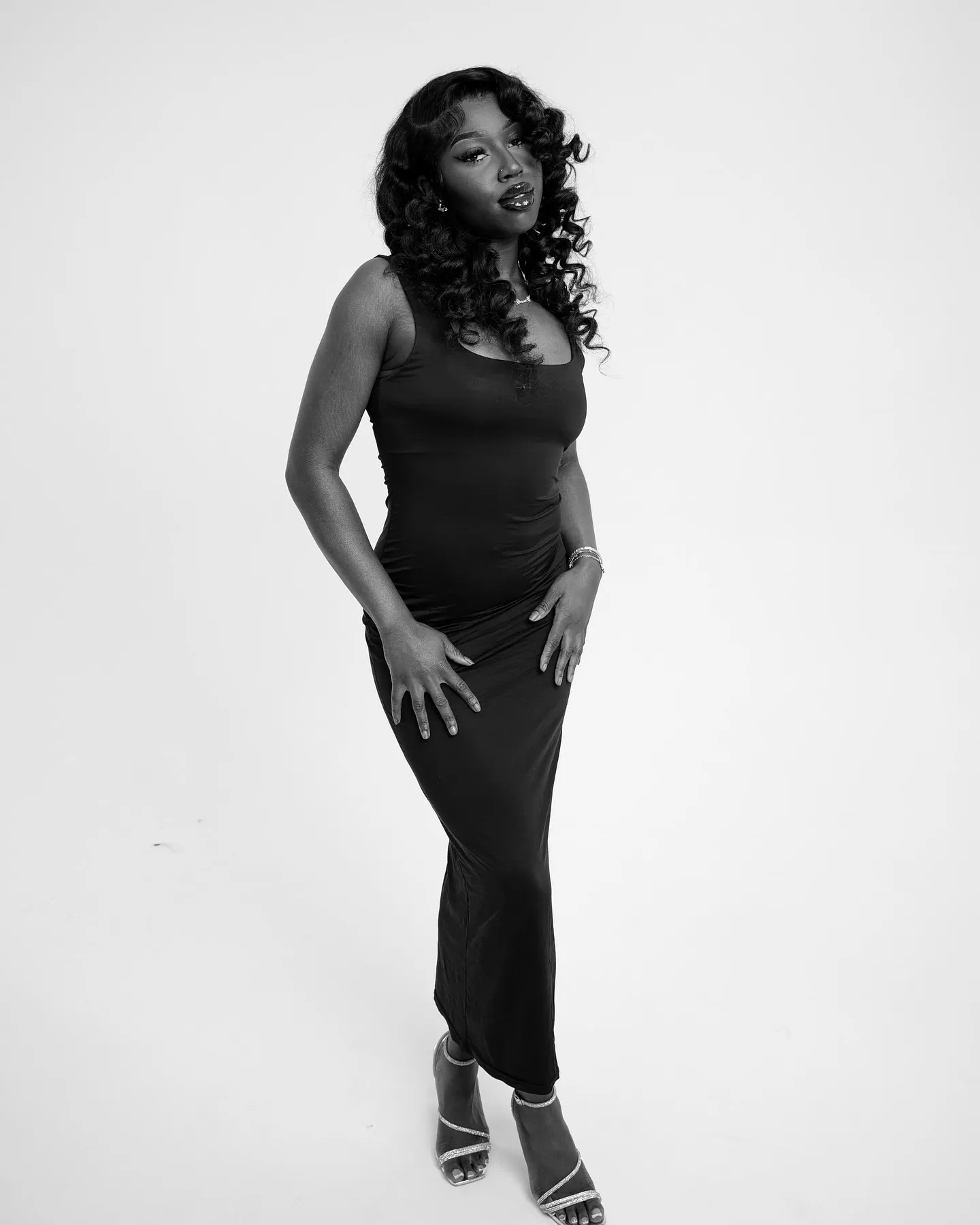 Black and white full-length shot of a woman in a long black dress and strappy heels, hands on hips, white backdrop