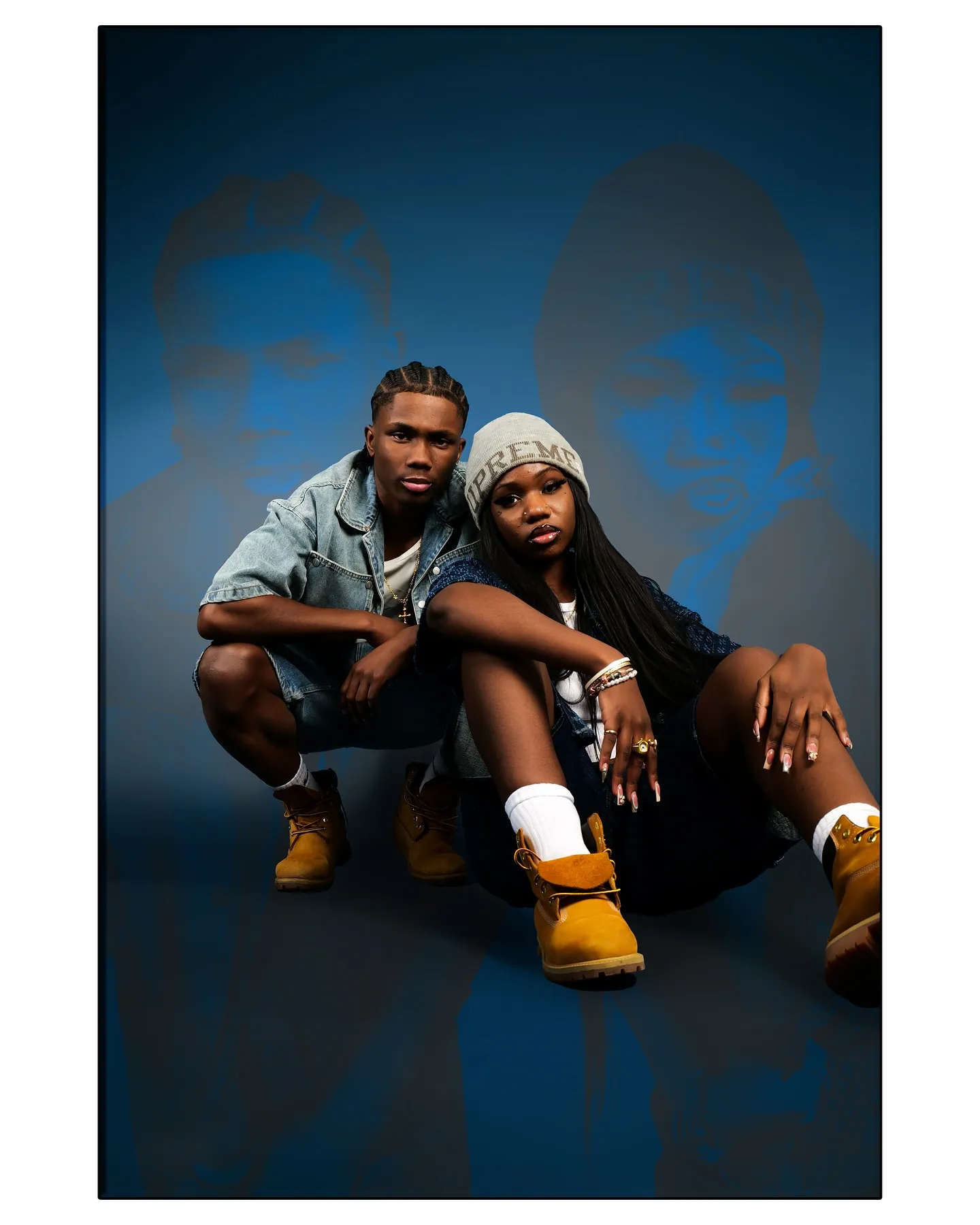 A man in a denim jacket and shorts crouching beside a woman in a grey beanie, against a blue backdrop with faded portraits