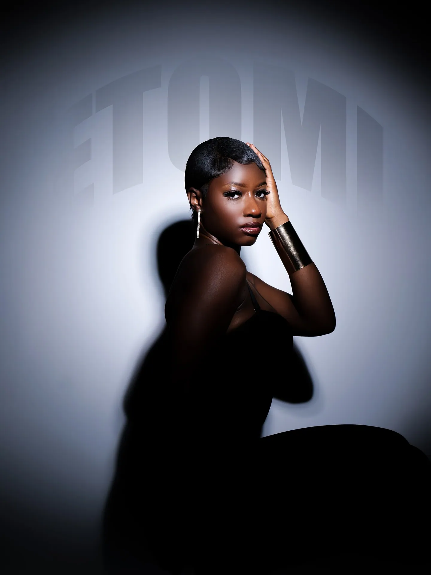 A woman with a short cropped hairstyle and gold cuff, hand on head, lit by a spotlight with ETOMI lettering behind her