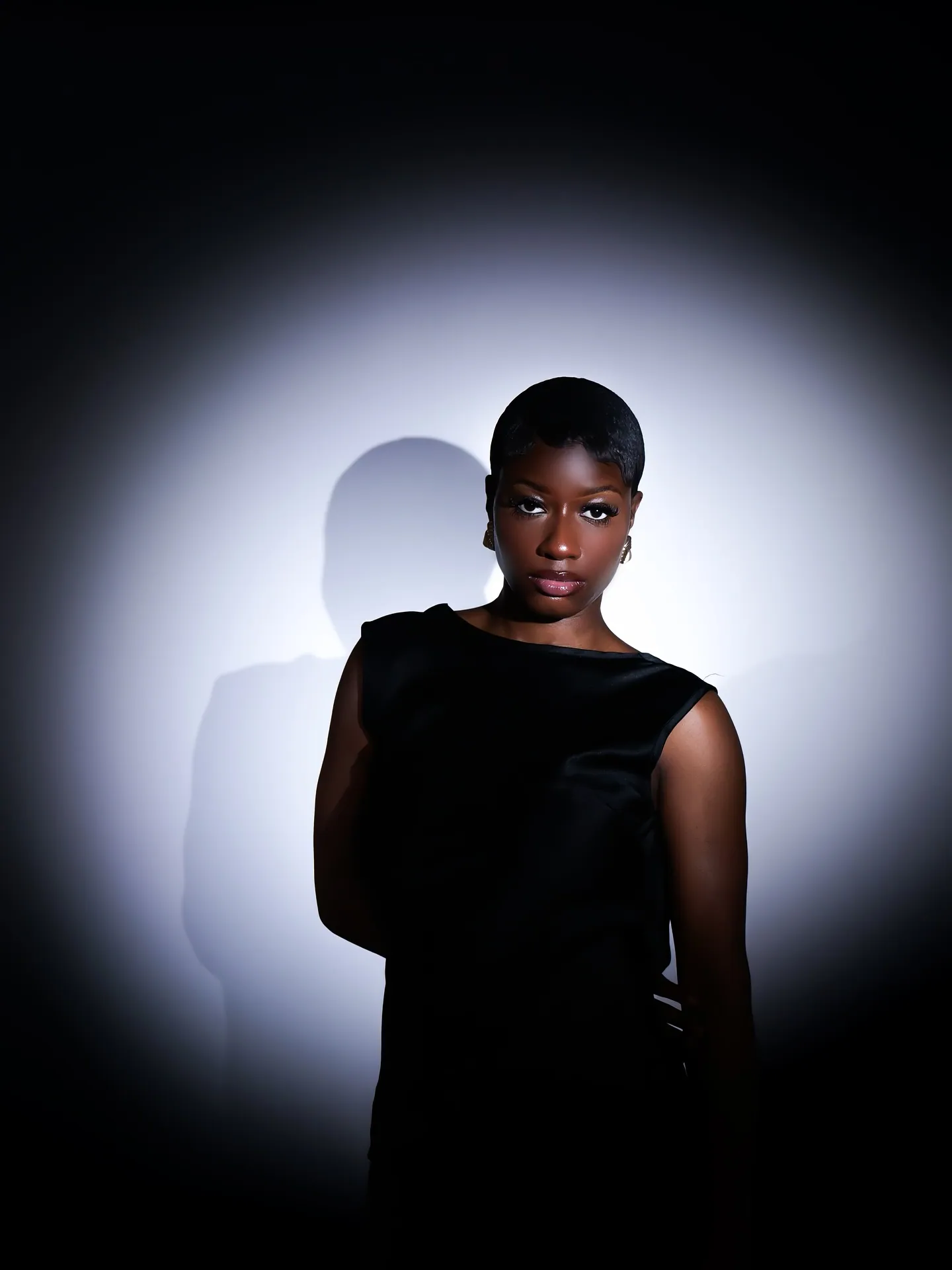A woman with short hair in a black sleeveless top standing in a circle of white light, her shadow on the wall behind