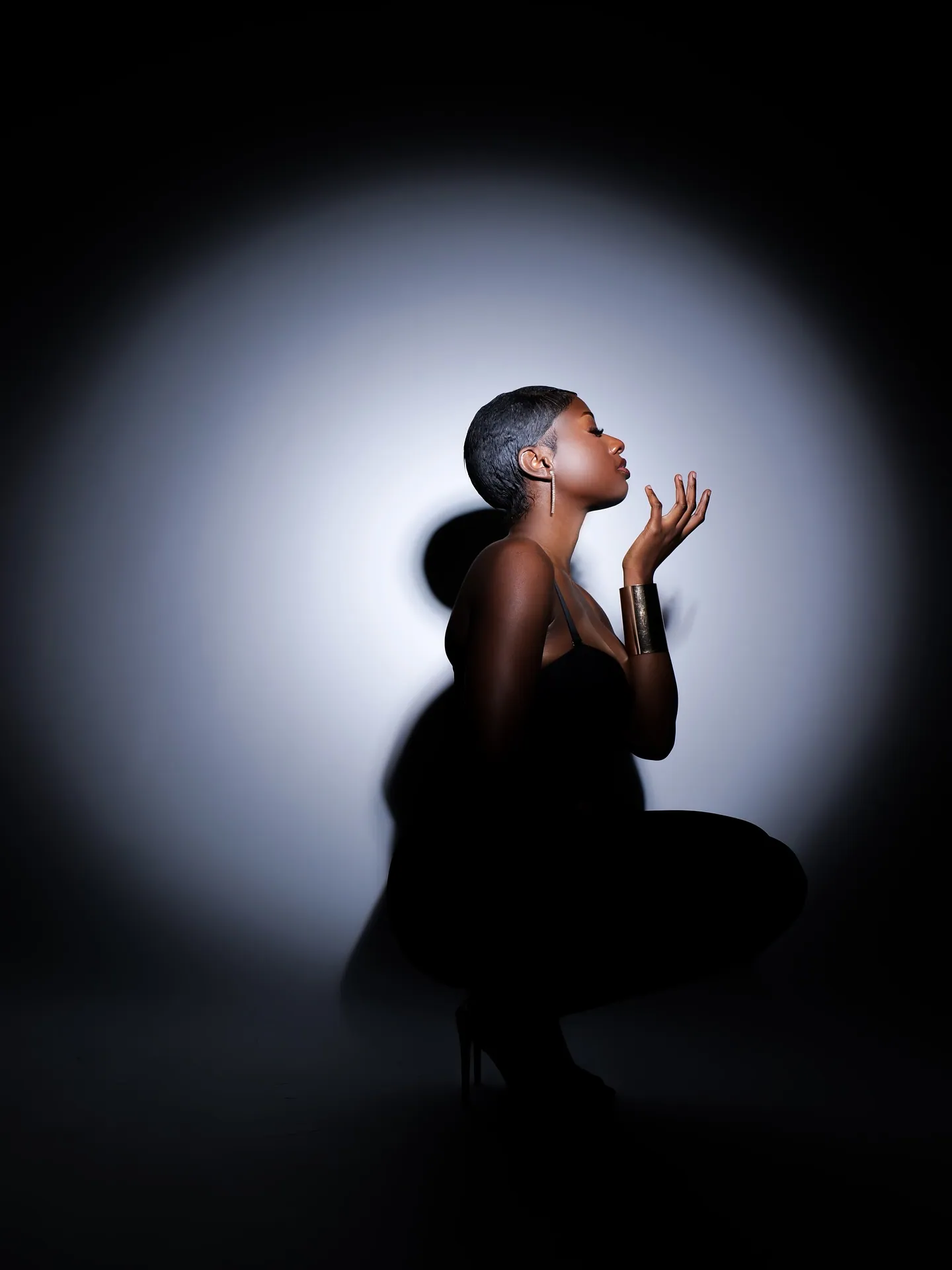 Side profile of a woman crouching in black with her hand raised near her lips, silhouetted in a circle of white light