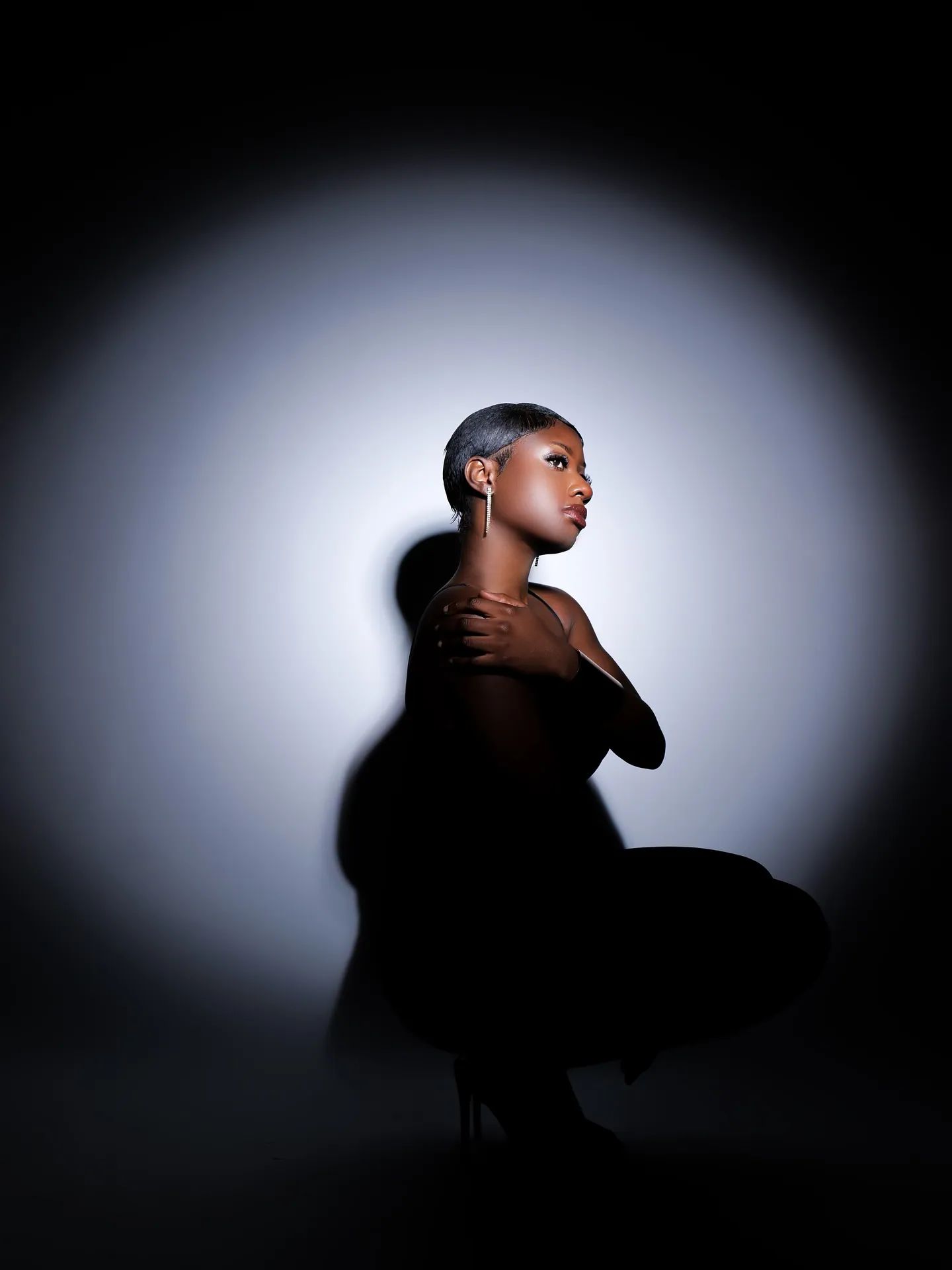A woman crouching in black, looking up with an arm across her chest, silhouetted in a circle of white light