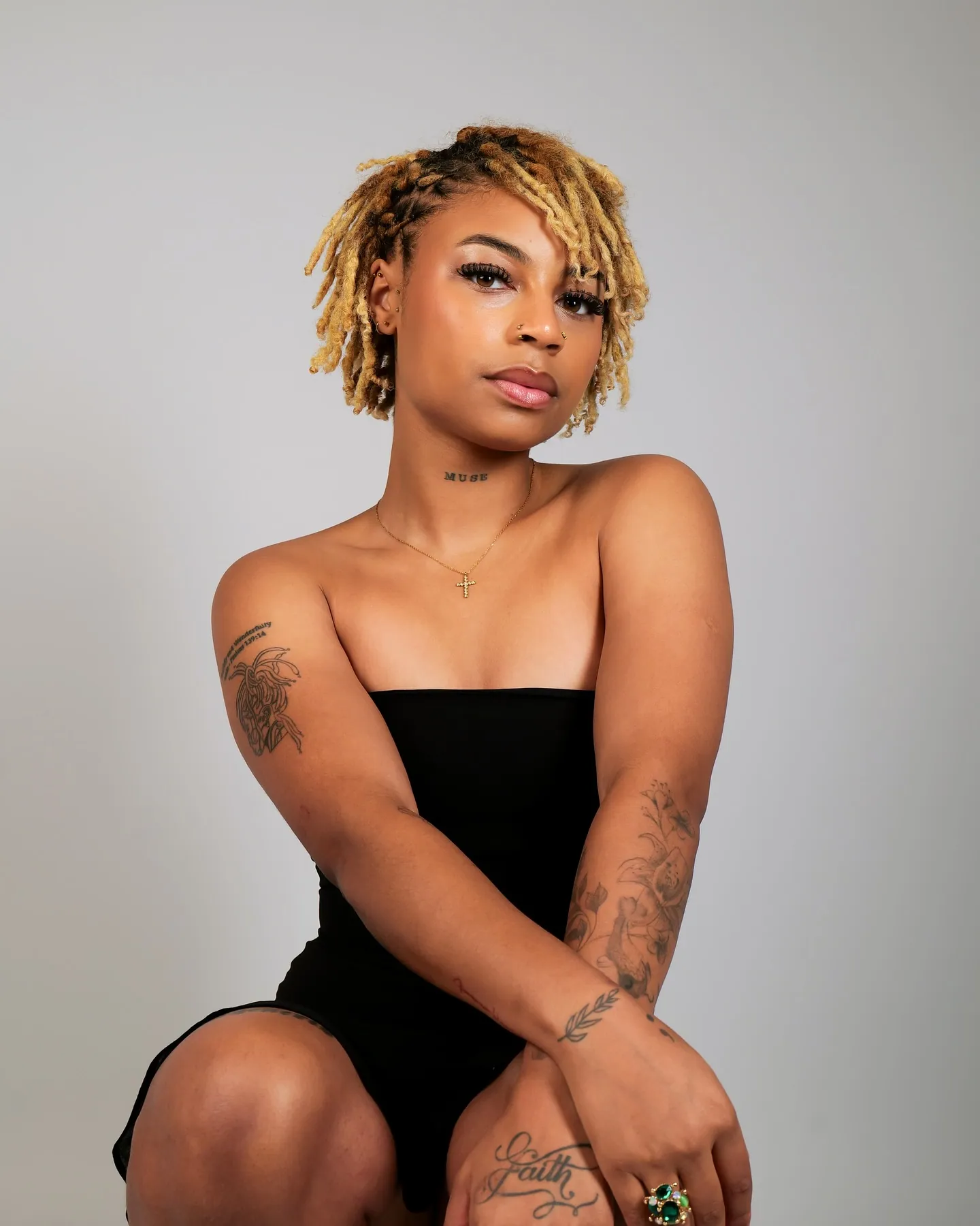 A woman with short blonde locs and tattoos in a black strapless dress, arms crossed over her knee, grey backdrop