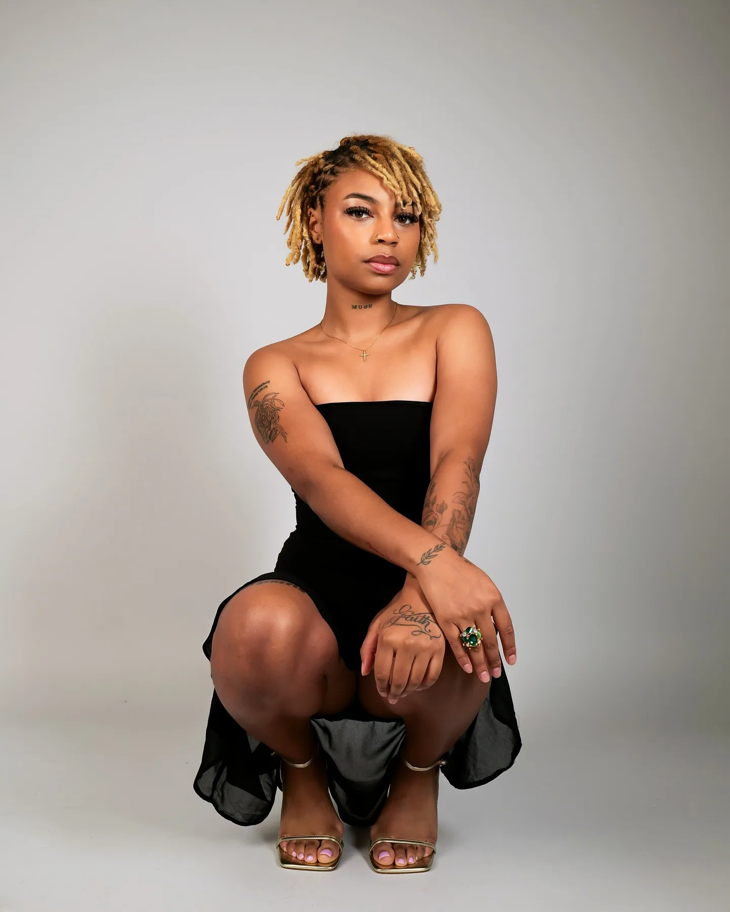 A woman with blonde locs and tattoos crouching in a black dress and gold strappy heels against a grey backdrop