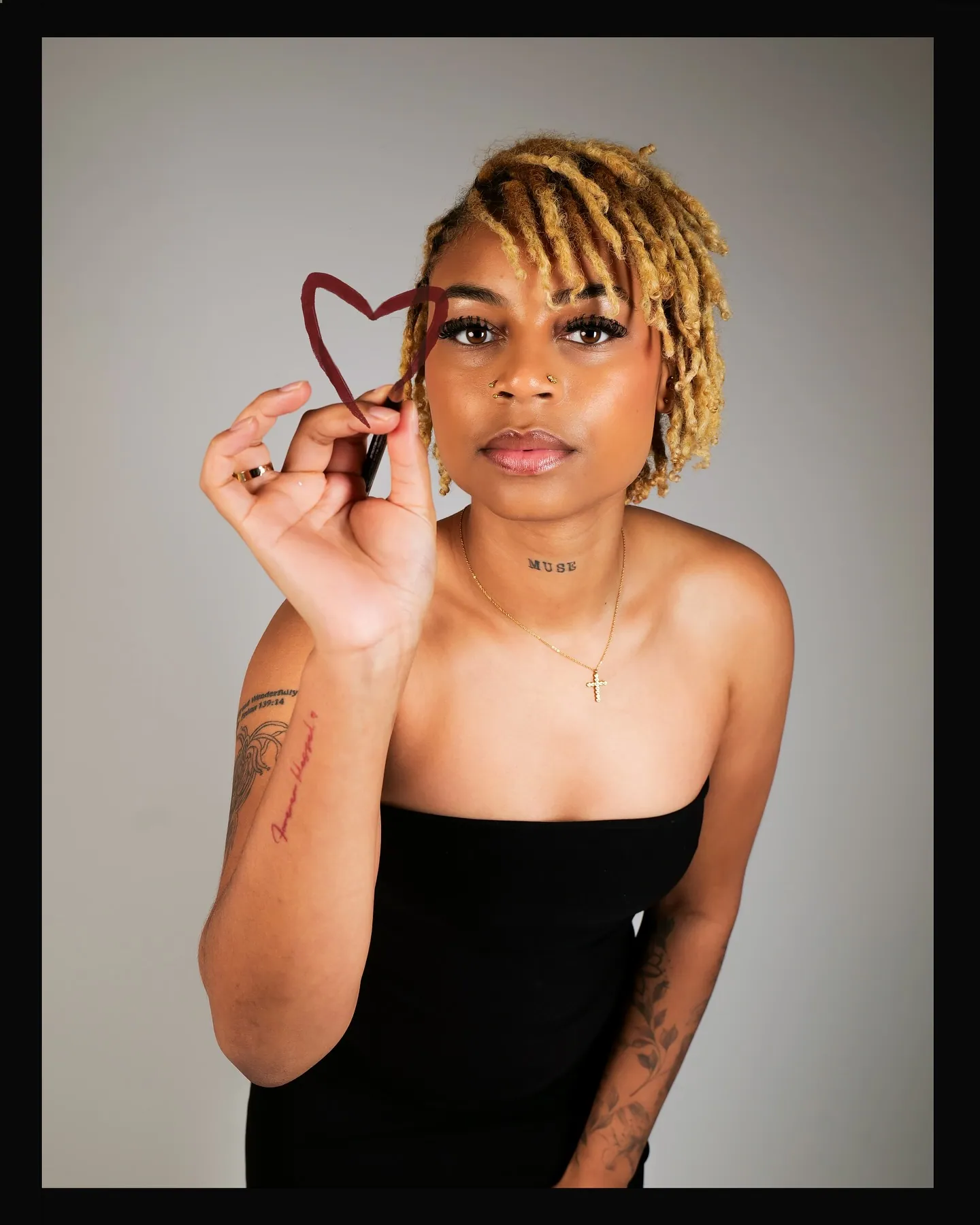 A woman with blonde locs and tattoos holding up a hand-drawn red heart, in a black strapless top, black border frame