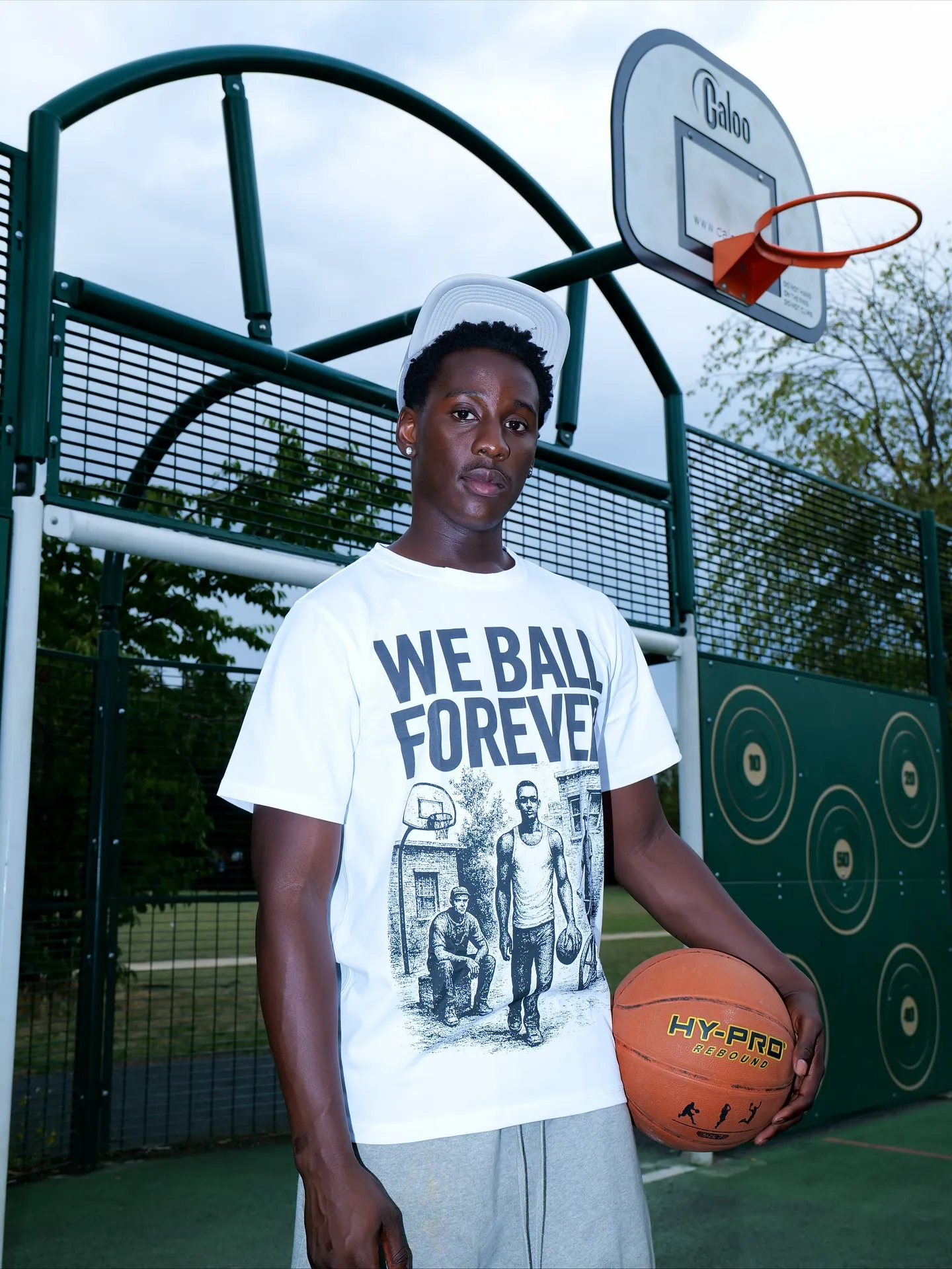 Man in a white WE BALL FOREVER t-shirt holding a basketball on an outdoor court, hoop behind him