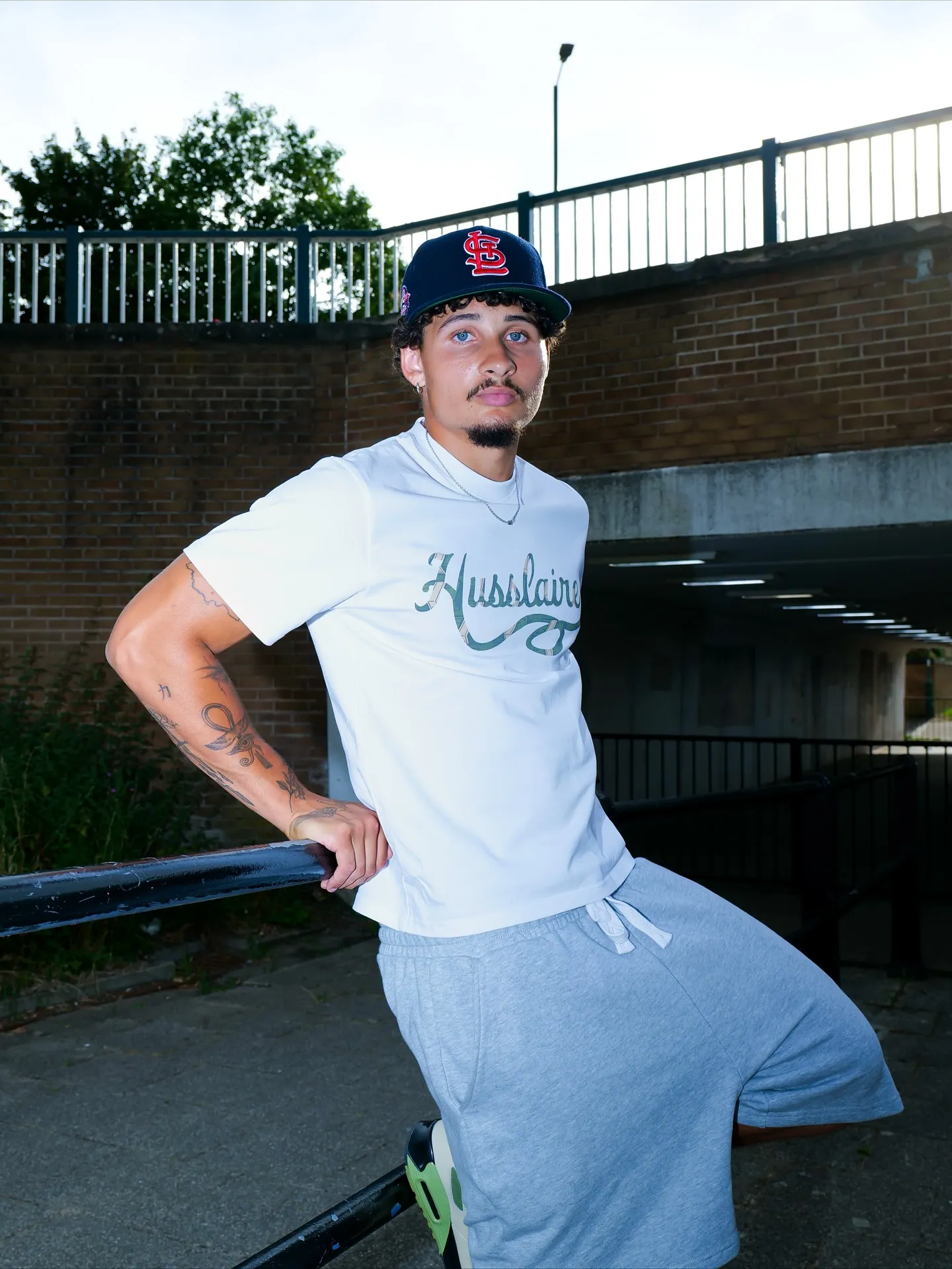 Man in a navy cap, white Husslaire t-shirt and grey shorts leaning on a railing in front of a brick underpass