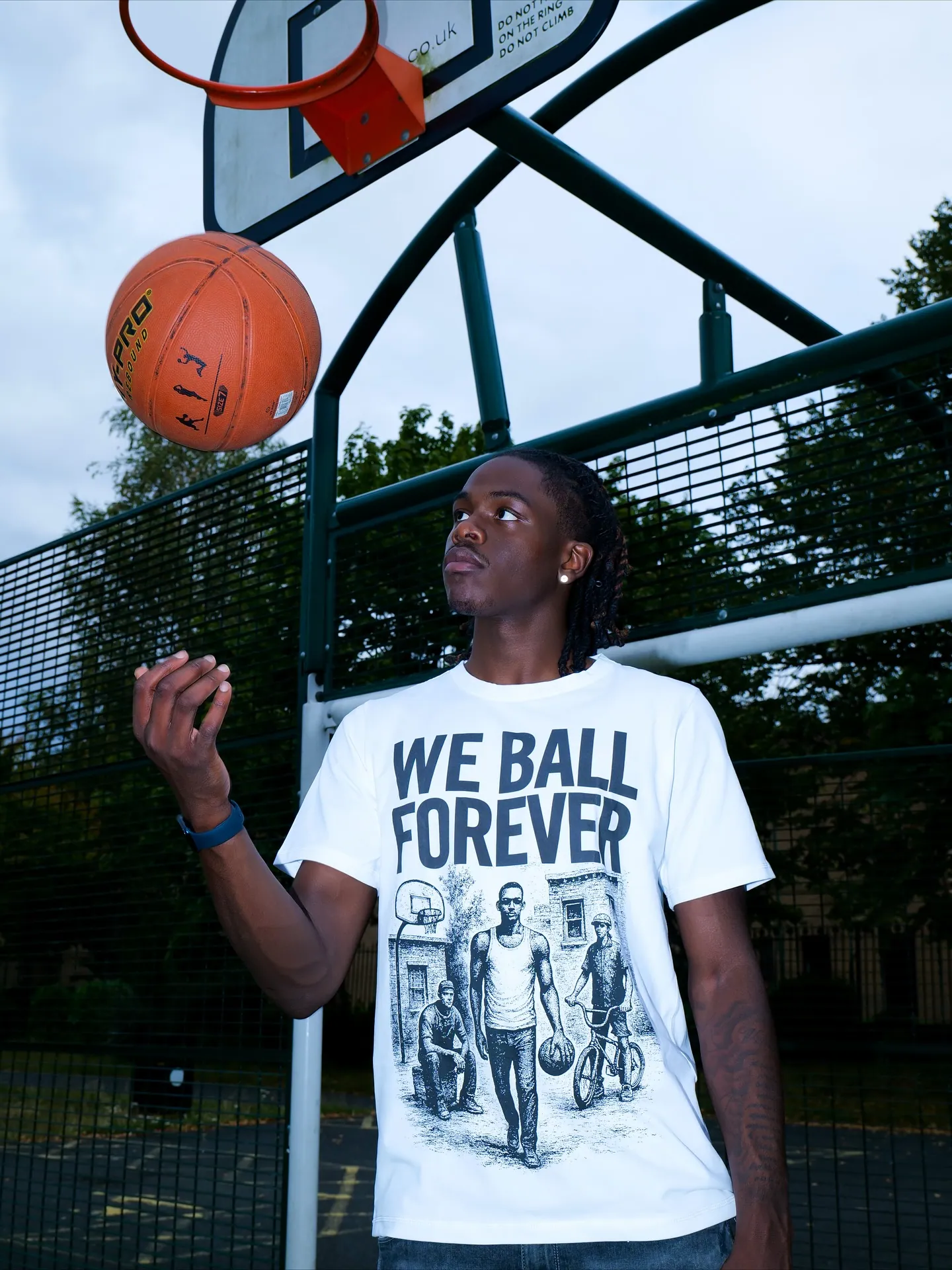 Man in a white WE BALL FOREVER t-shirt looking up at a basketball spinning near a hoop and fence
