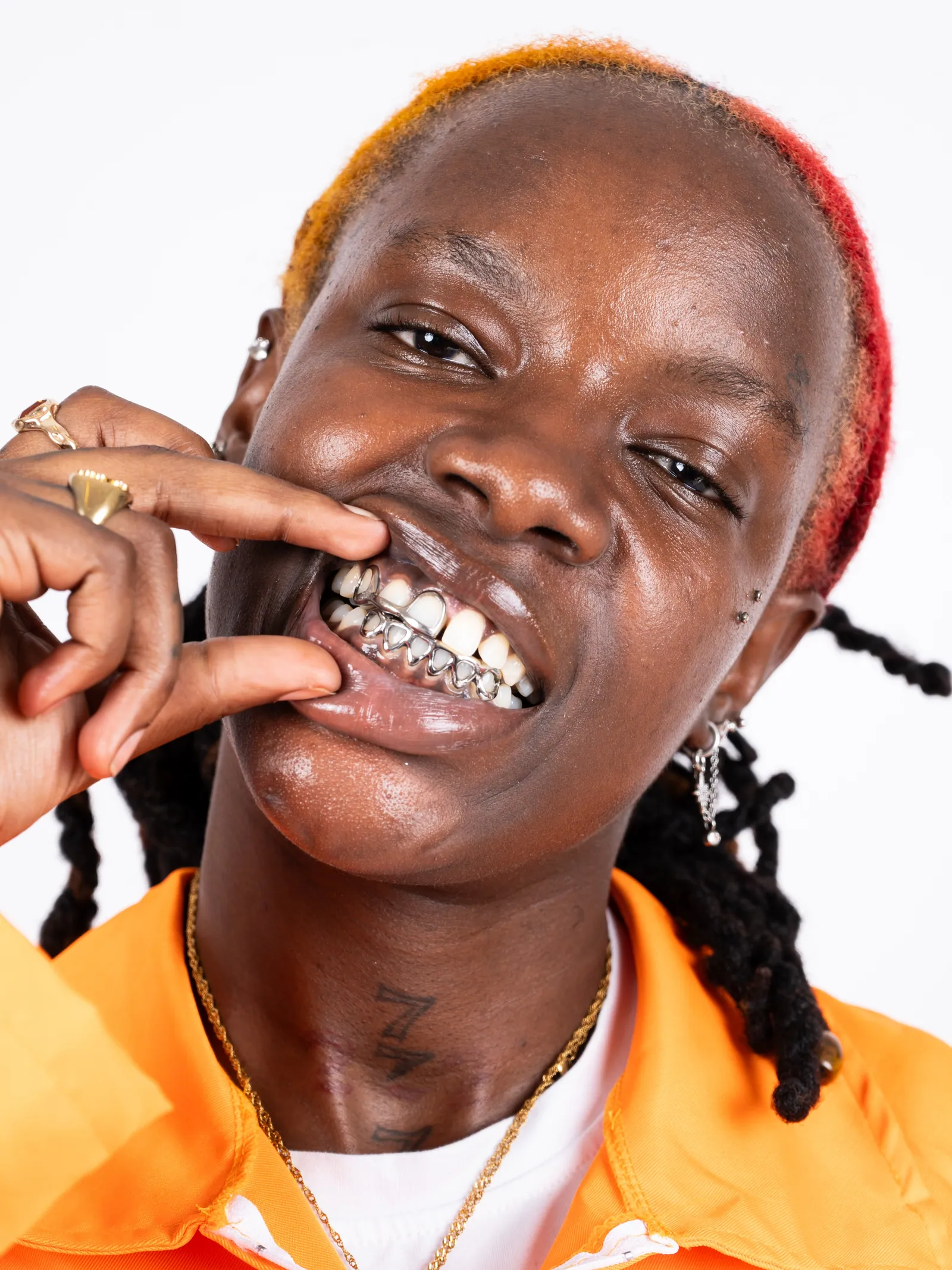 A smiling person in an orange jumpsuit with silver grills, biting a finger, red and orange dyed hair, white backdrop