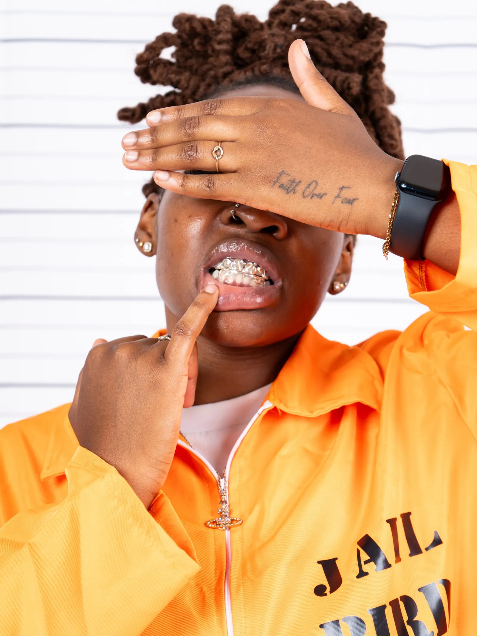 A person in an orange JAIL BIRD jumpsuit covering their eyes with a hand tattooed Faith over Fear, grills on show