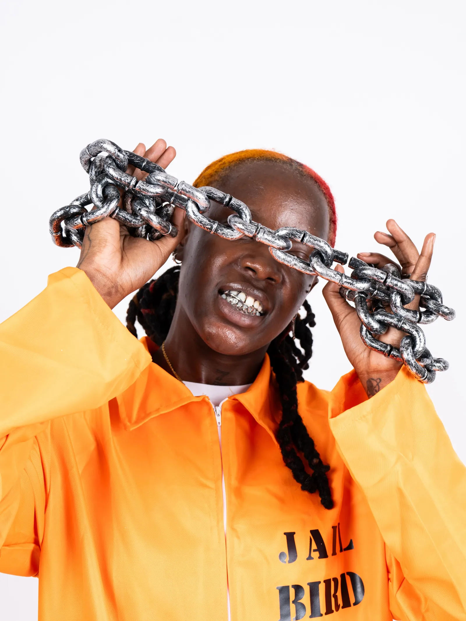 A person in an orange JAIL BIRD jumpsuit holding a heavy silver chain across their face, white background