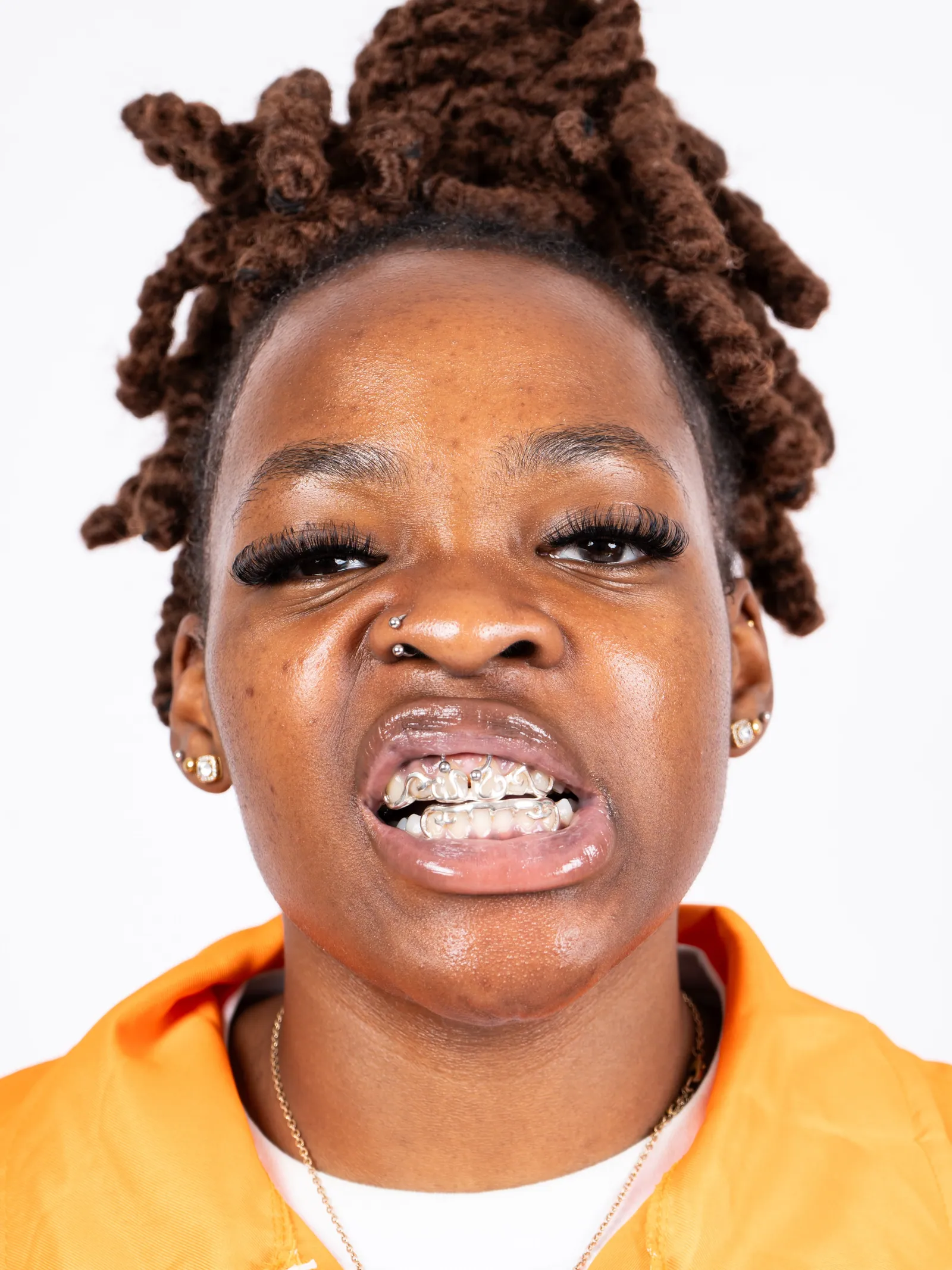 Close-up of a person with twisted locs and jewelled grills pulling a stern face, orange collar visible, white backdrop