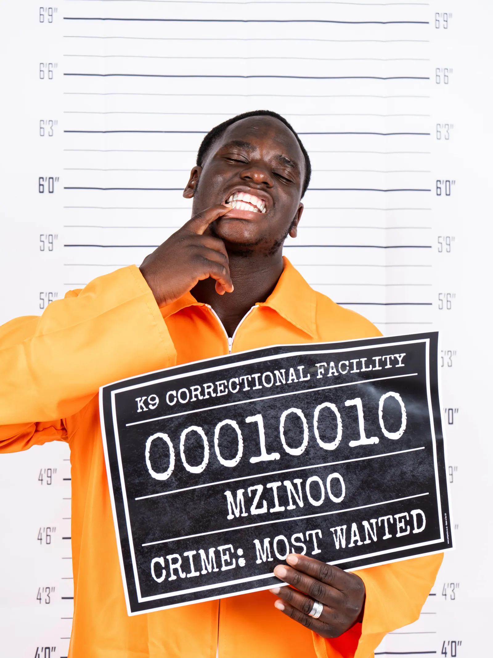 Mugshot-style portrait of a smiling man in an orange jumpsuit holding a K9 Correctional Facility board reading MZINOO