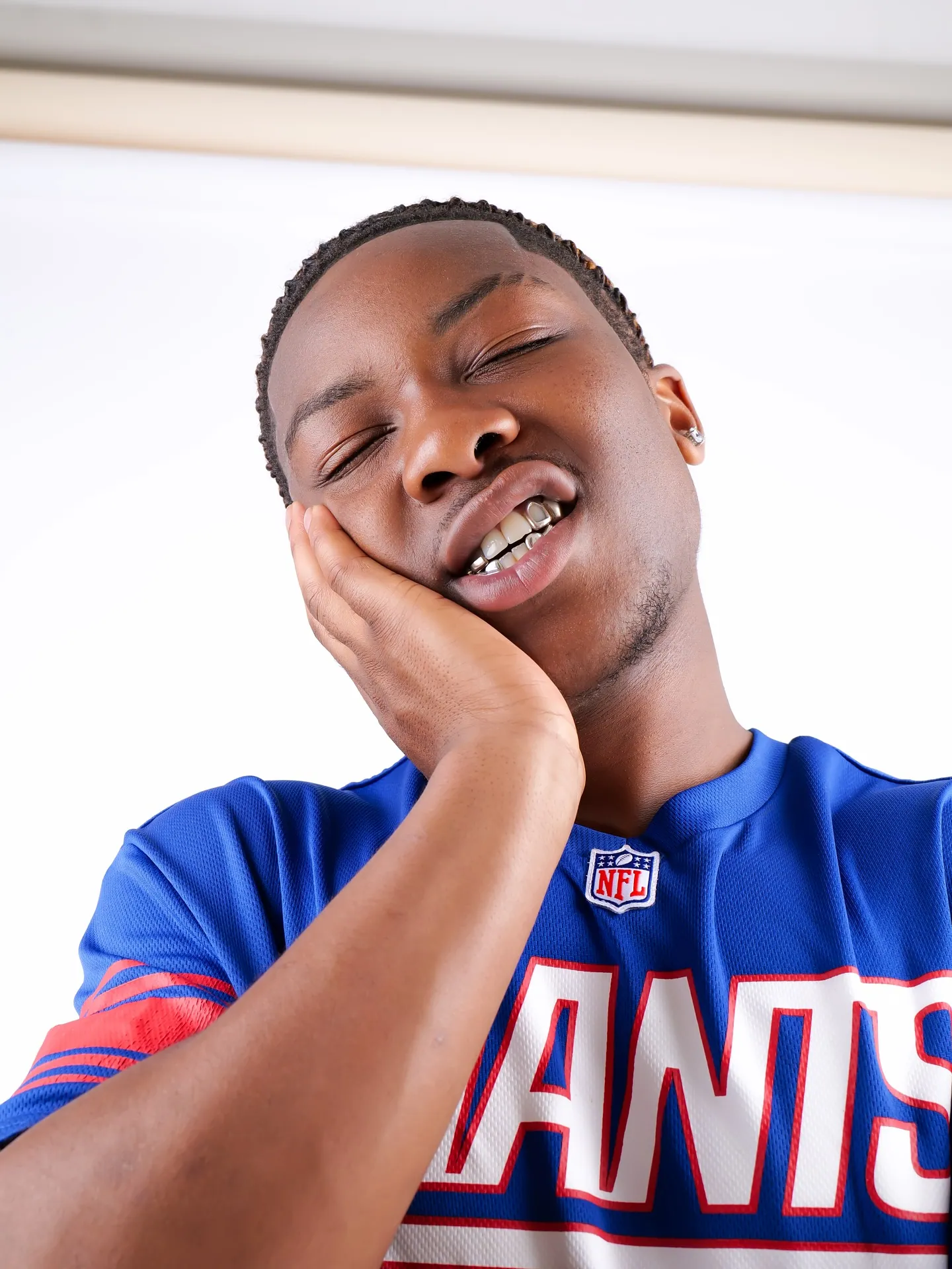 A man in a blue Giants jersey with eyes closed and cheek resting on his hand, showing a grill, seen from below