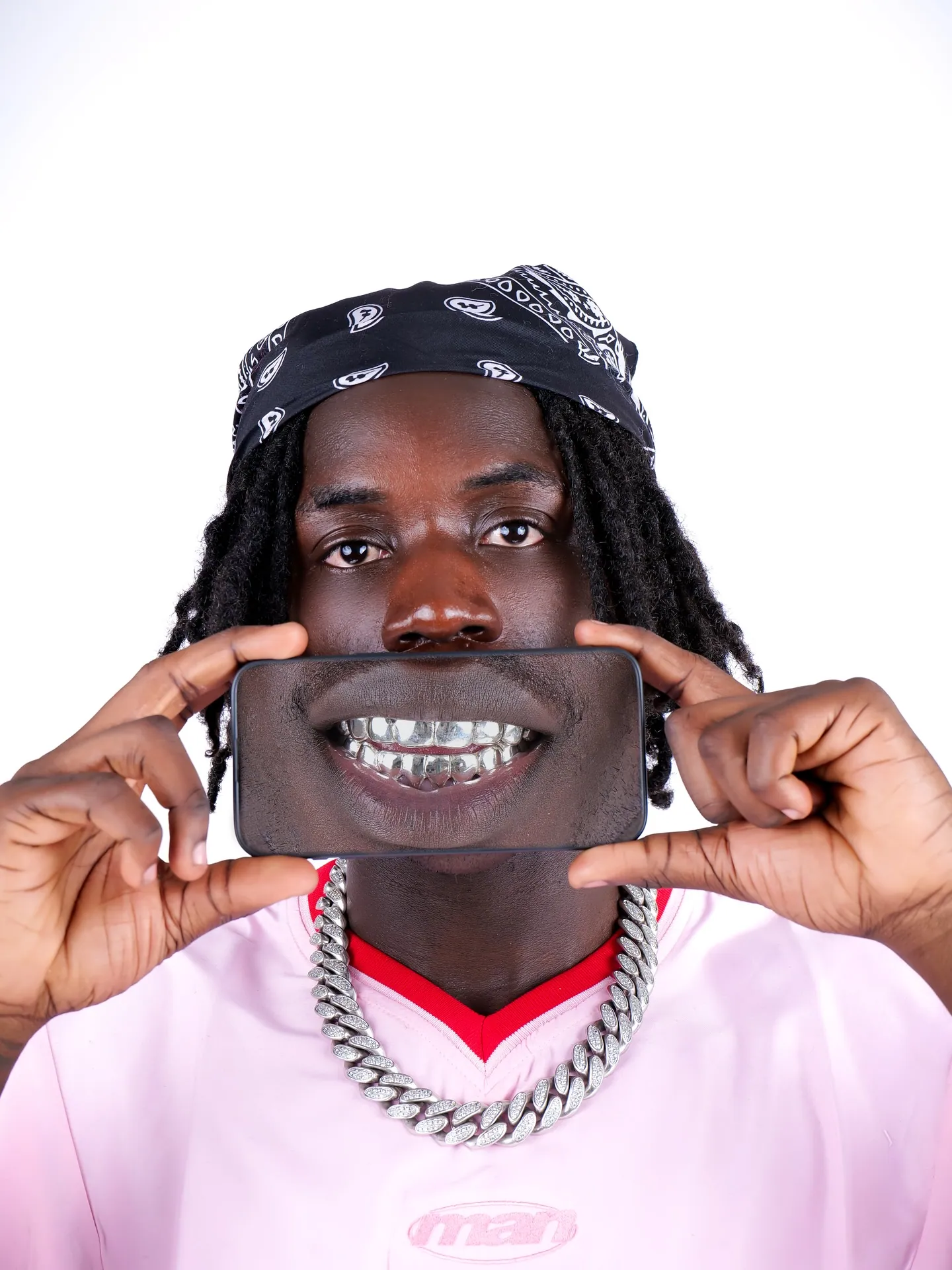 A man in a bandana, chain and pink top holding a phone screen showing a grill smile over his mouth, white backdrop