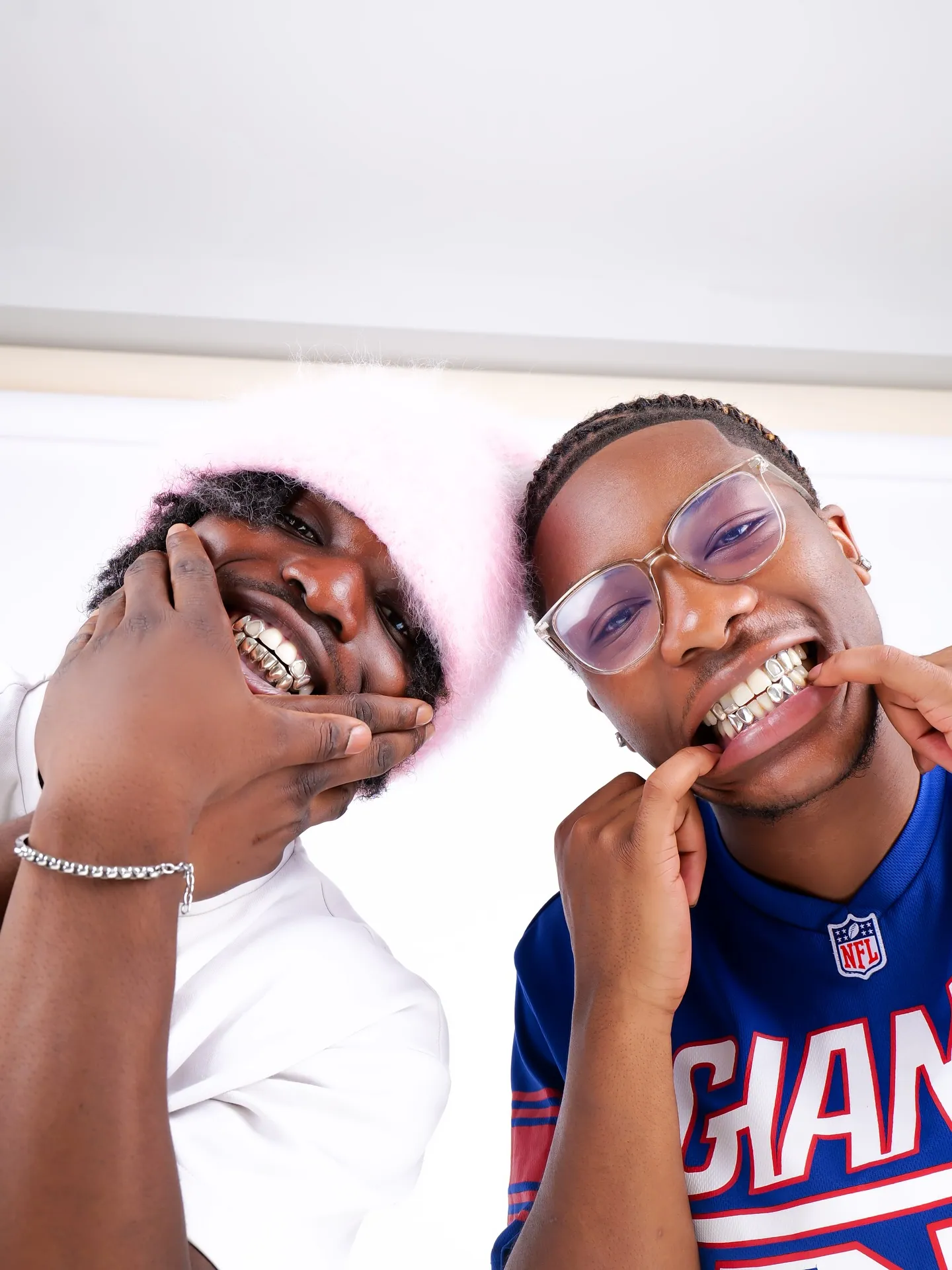 Two smiling men showing grills, one in a pink fluffy hat and white tee, one in clear glasses and a Giants jersey