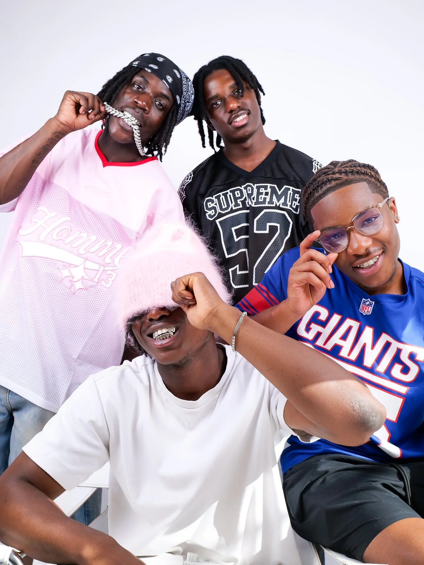 Four friends posed together in sports jerseys, a pink fluffy hat and a bandana, one holding a chain in his mouth