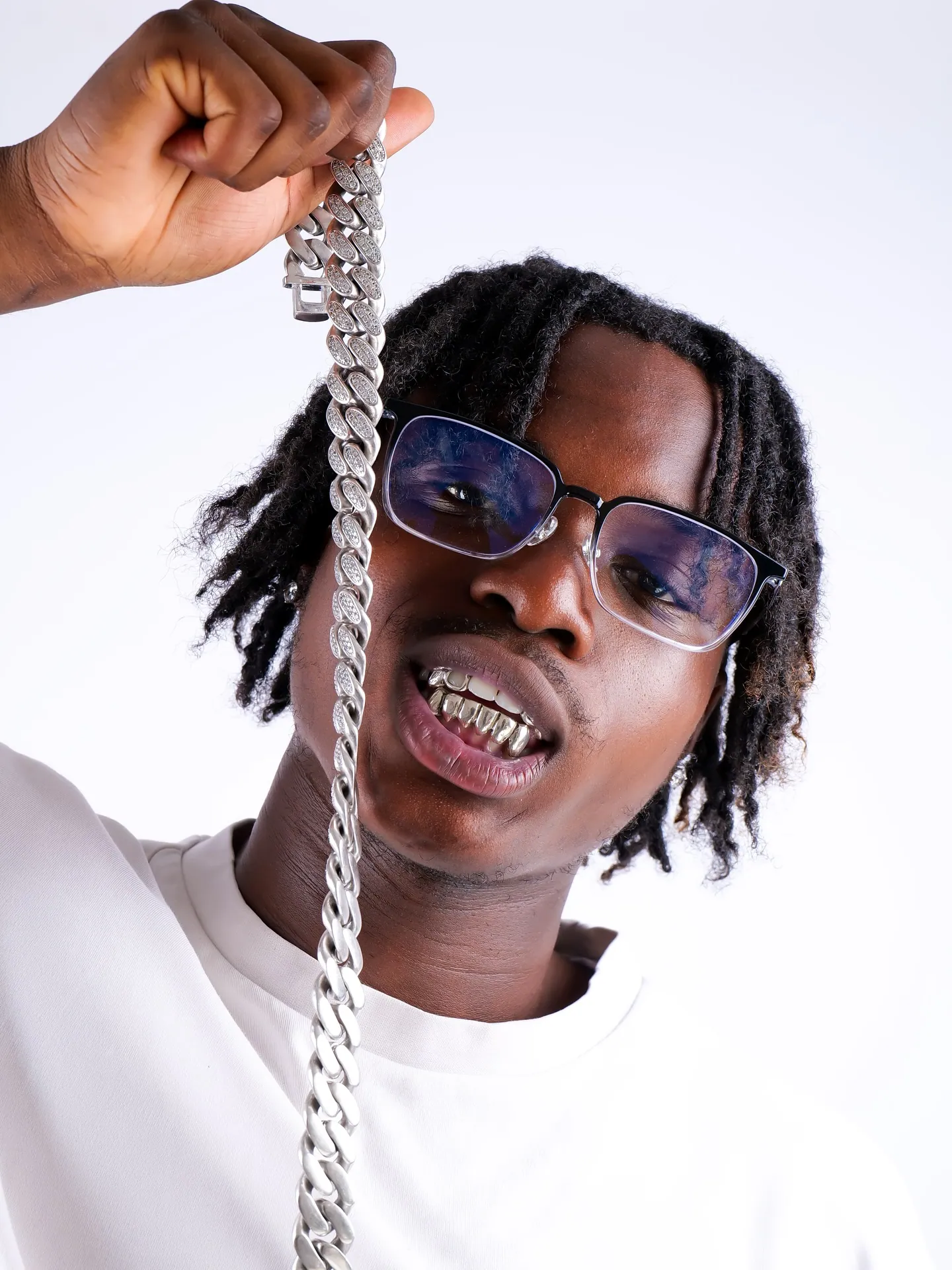 A man in tinted glasses and white tee holding up a large silver Cuban chain while showing a grill, white backdrop