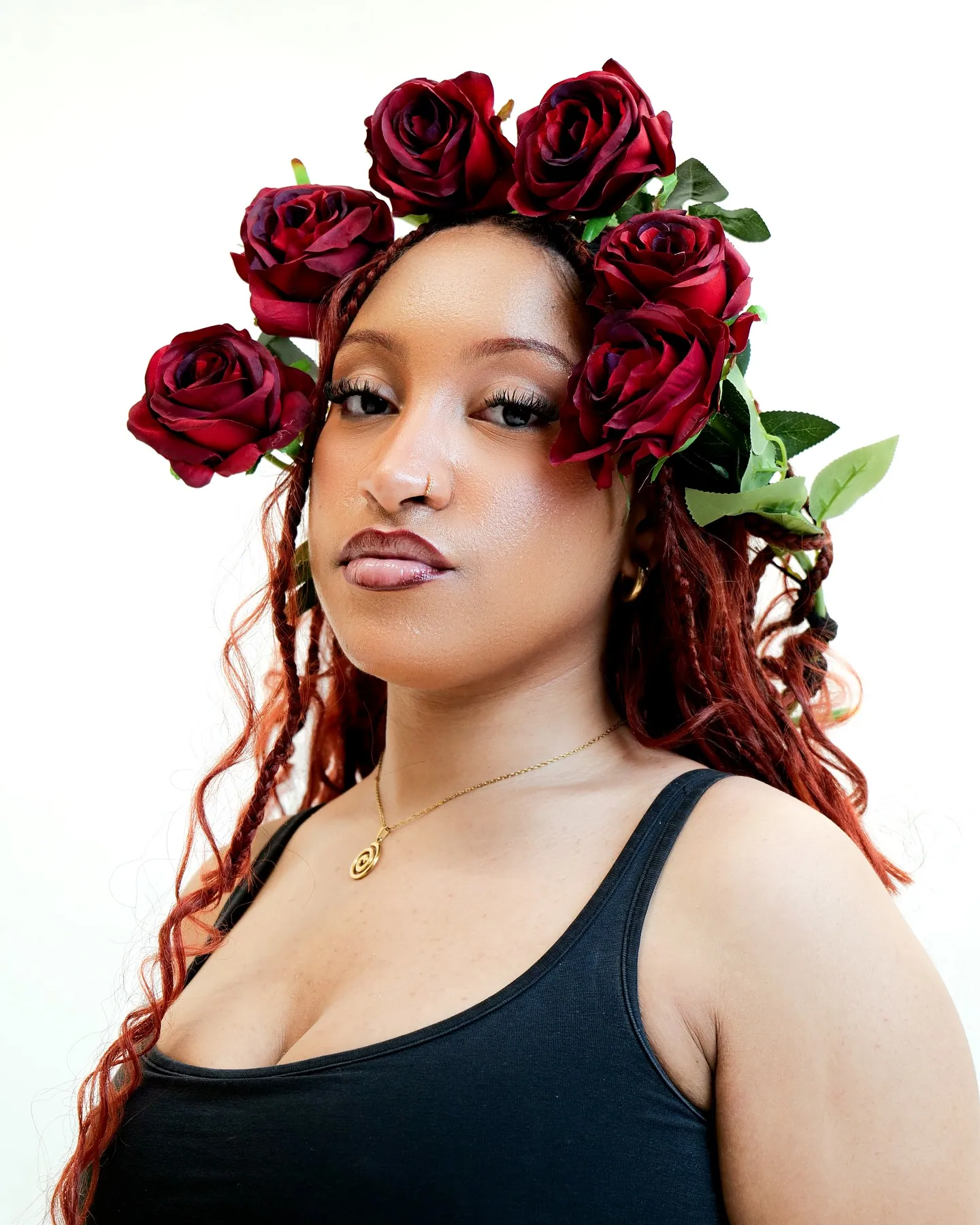 Portrait of a woman with red braids wearing a crown of dark red roses, black vest and spiral gold pendant, white backdrop