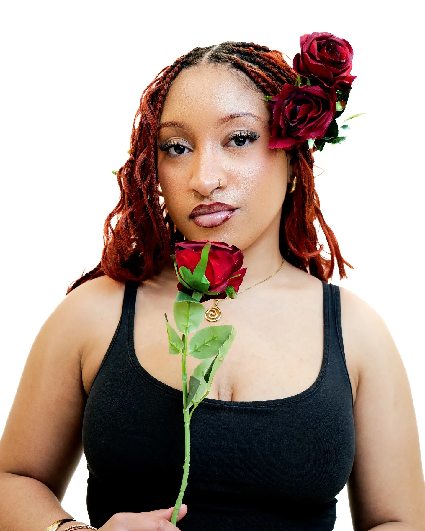 A woman with red braids holding a red rose below her chin, red roses pinned in her hair, black vest, white backdrop