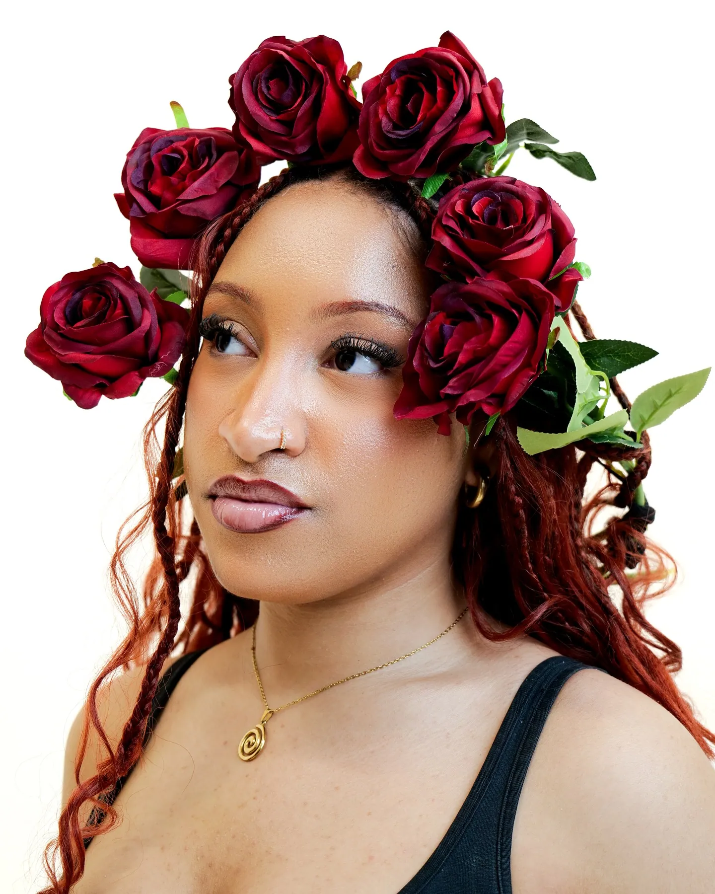 Side-on portrait of a woman with red braids and a crown of red roses, gold spiral pendant, white background
