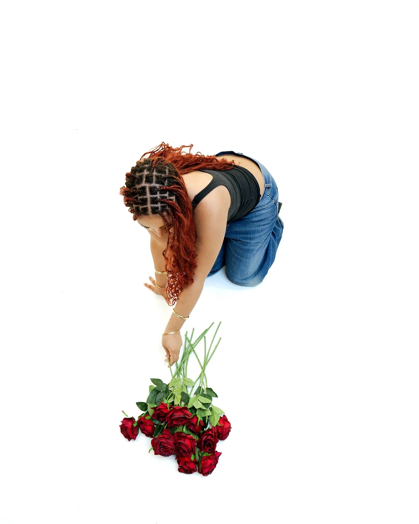 High-angle view of a woman kneeling in blue jeans and a black top, reaching down to a bunch of red roses, white floor
