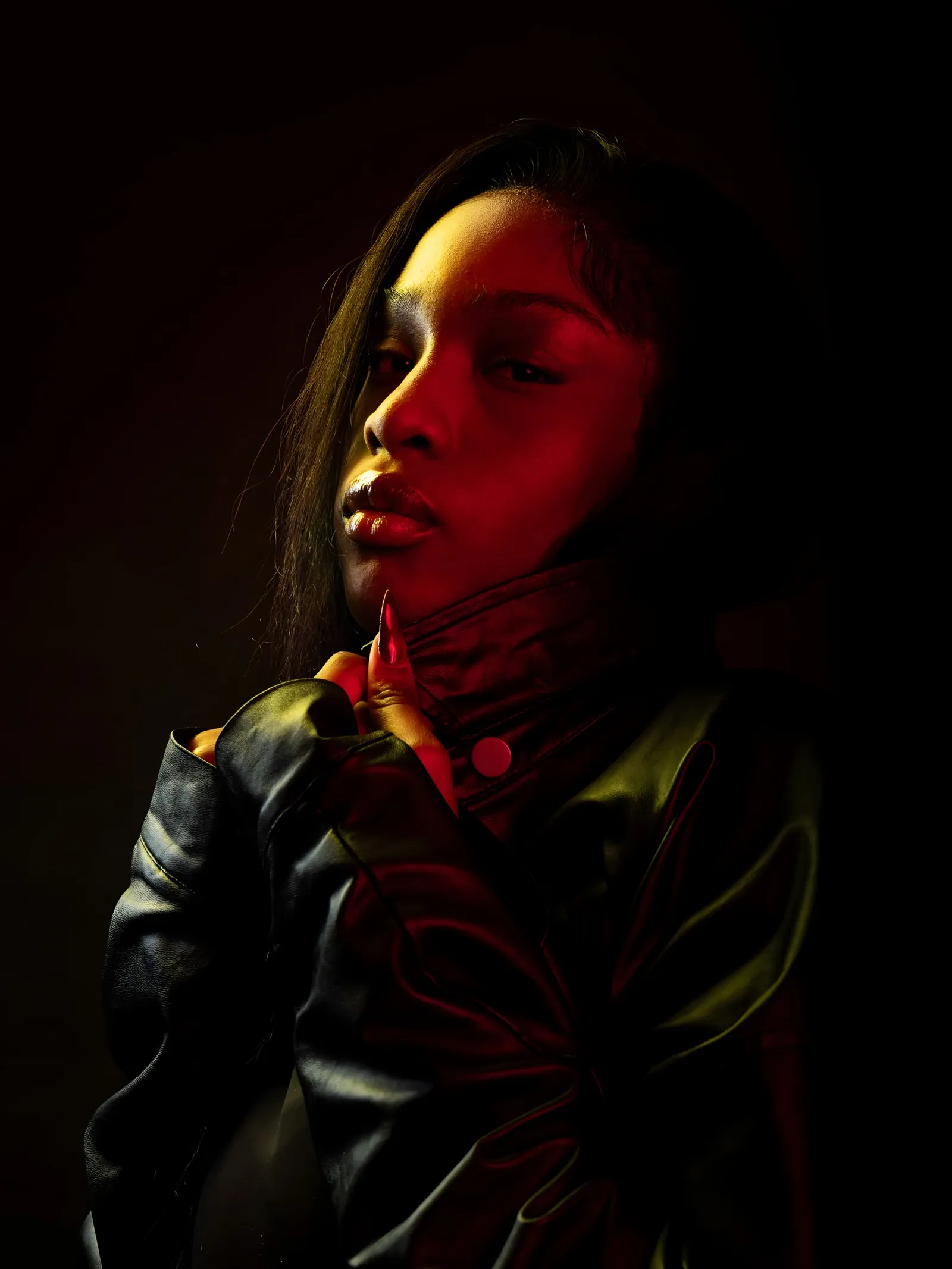 Model in a high-collared leather jacket with a finger at her chin, glancing sideways in low red and amber light
